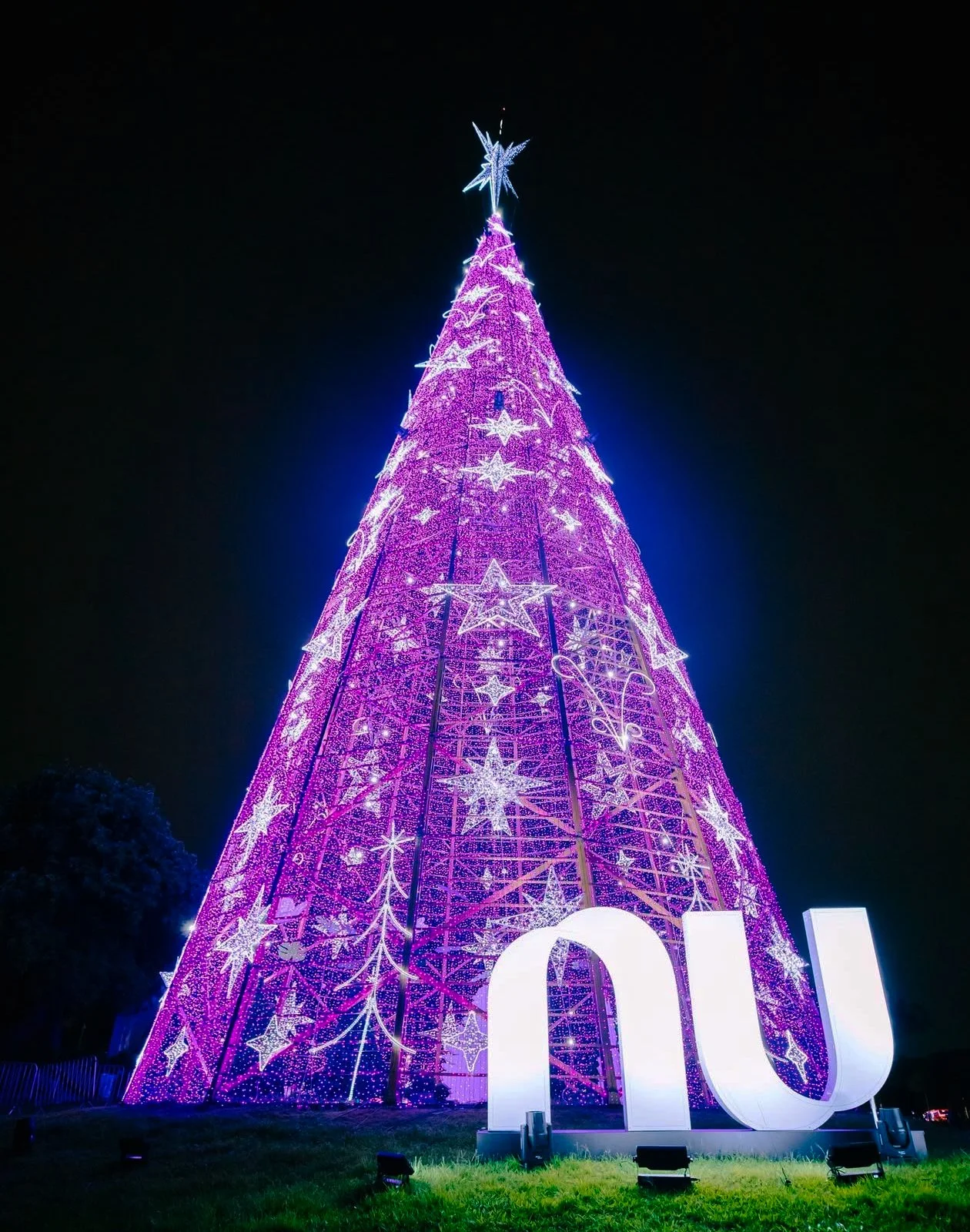 Árvore de Natal iluminada de rosa com estrelas brancas e enfeites luminosos, ao lado há uma estrutura de letras brancas formando a palavra 'Noel', tudo contra um céu noturno.