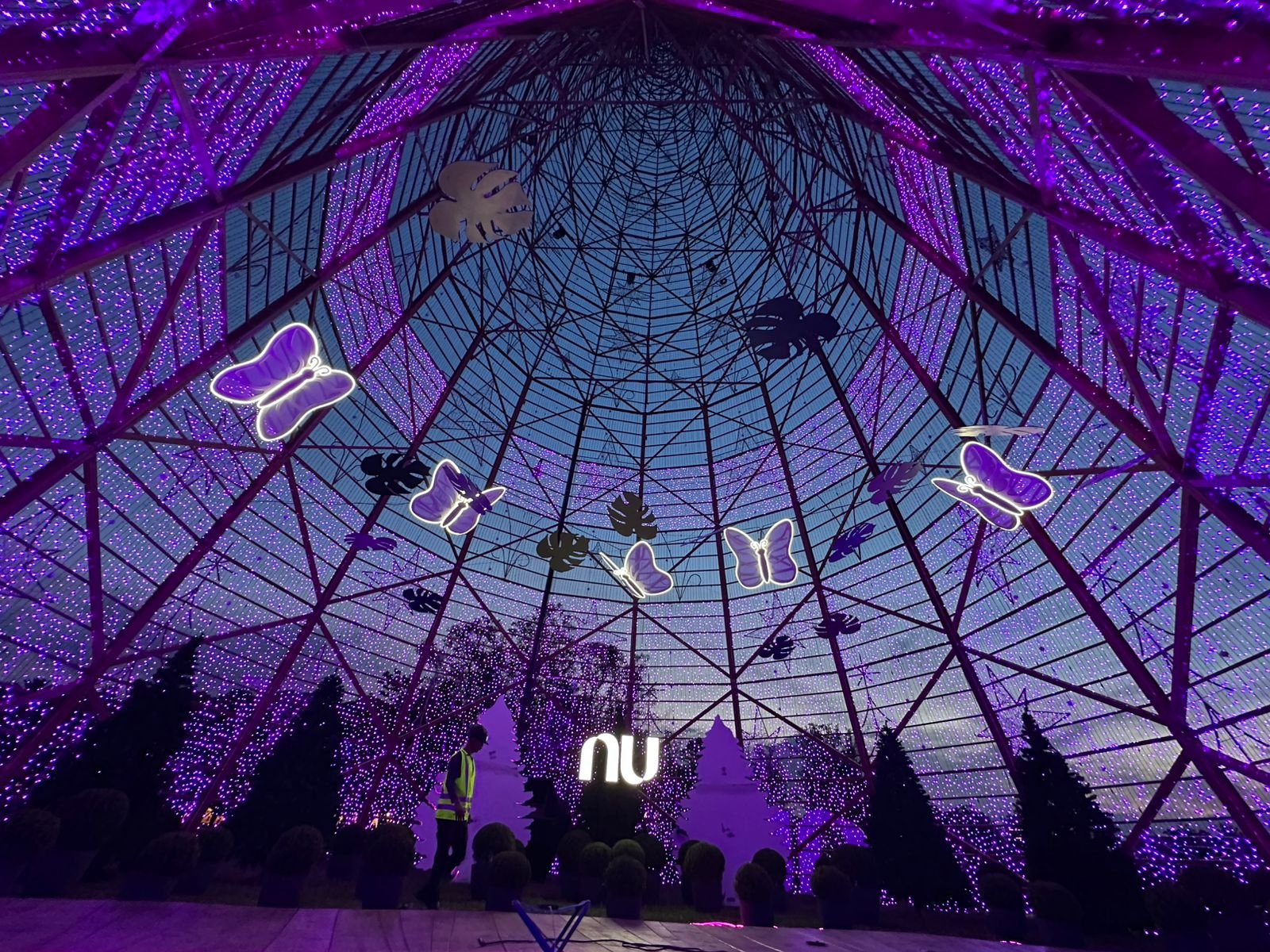 Estrutura de ferro com iluminação roxa, decorada com borboletas e folhas e um céu ao fundo, em um evento ou atração noturna.