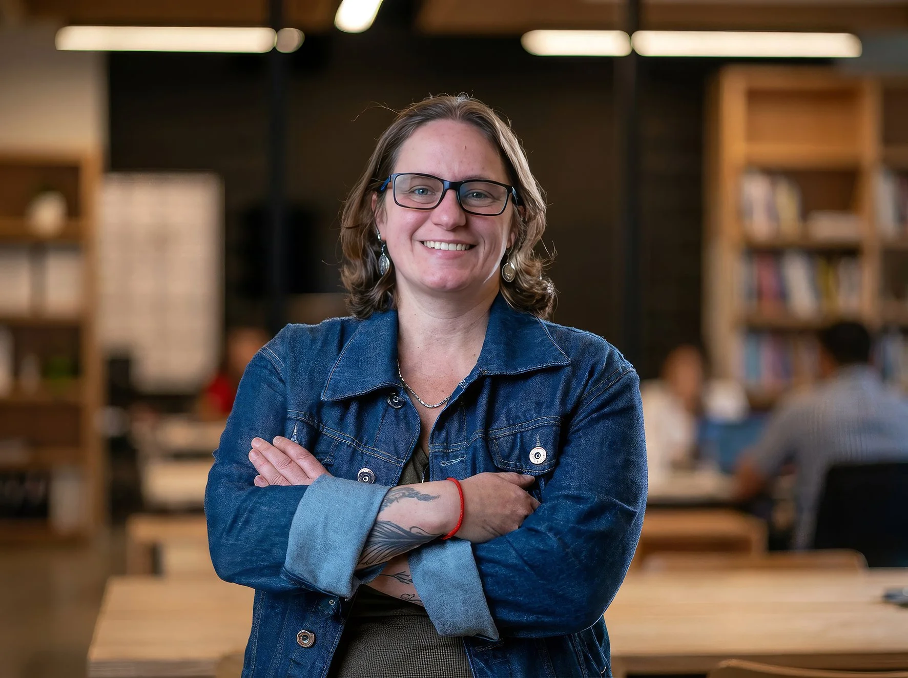 Mulher sorridente com óculos e cabelo cacheado preso, vestindo jaqueta de ganga, em ambiente com biblioteca ao fundo, braços cruzados e expressão confiante.
