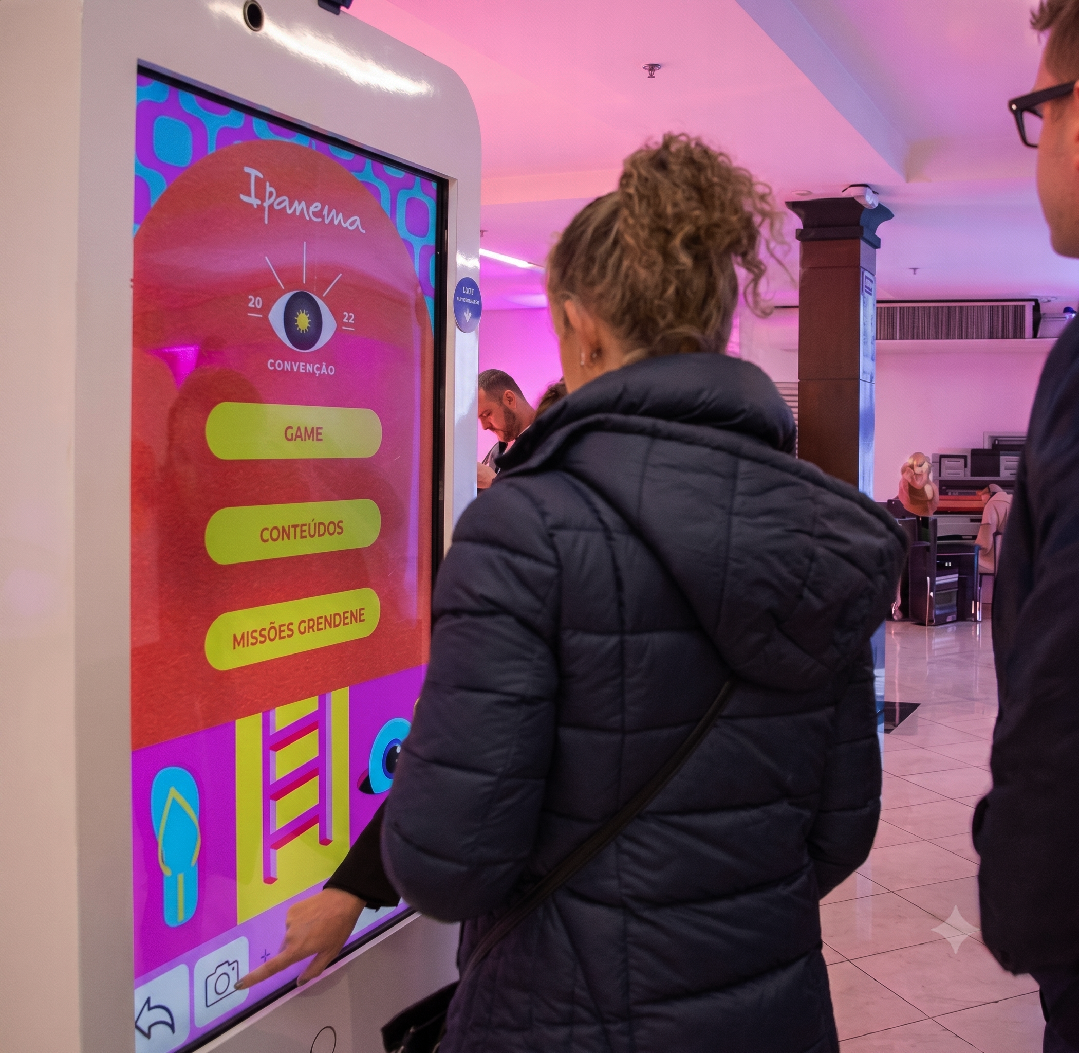 Pessoa com cabelo cacheado e óculos tocando na tela de um quiosque digital com tema colorido e texto em português, em ambiente de shopping.
