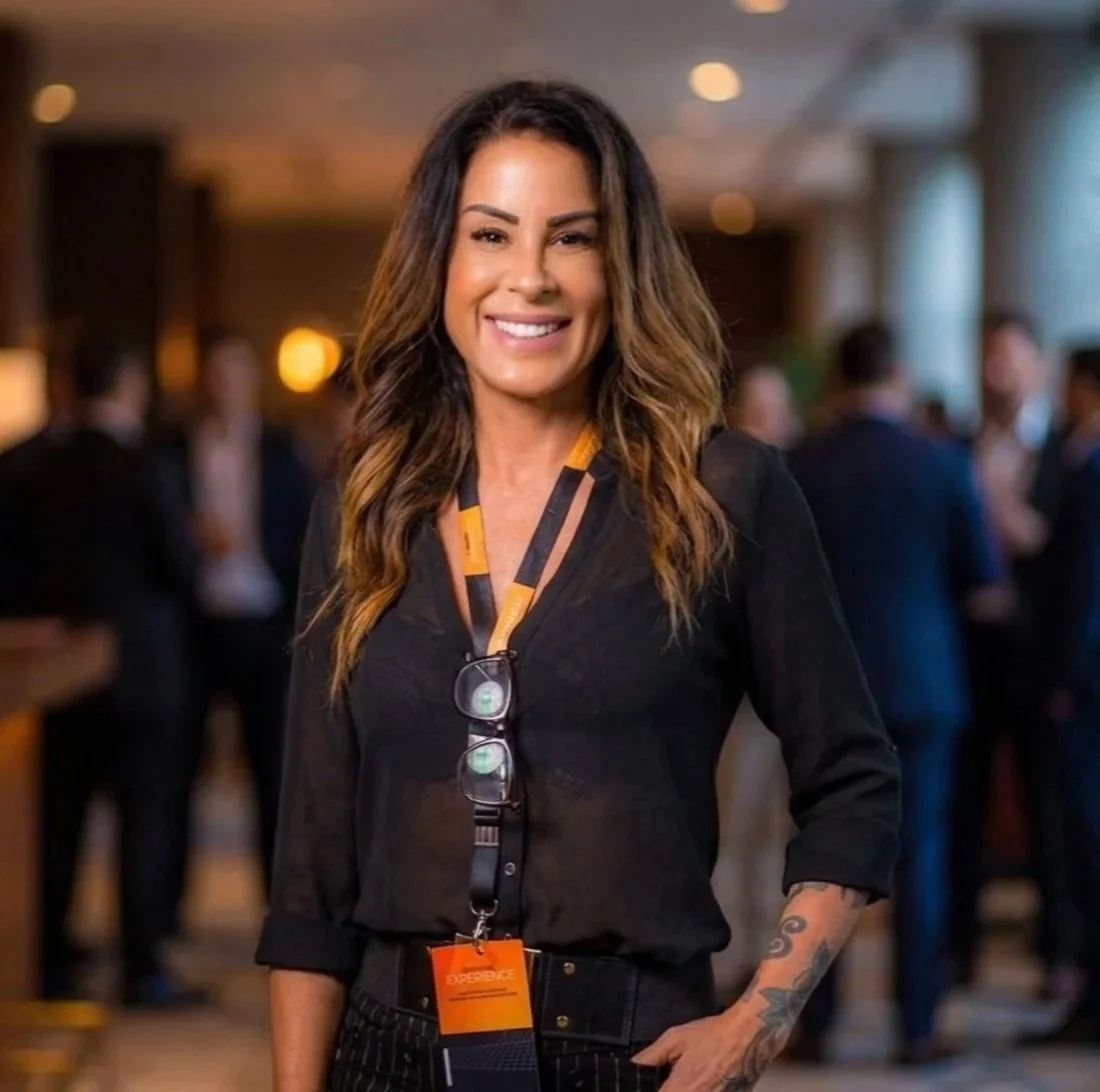 Mulher sorridente com cabelo castanho e ondulado, usando camiseta preta, óculos pendurado no pescoço, com uma experiência de evento no pescoço, em ambiente com pessoas ao fundo em um espaço interno bem iluminado.