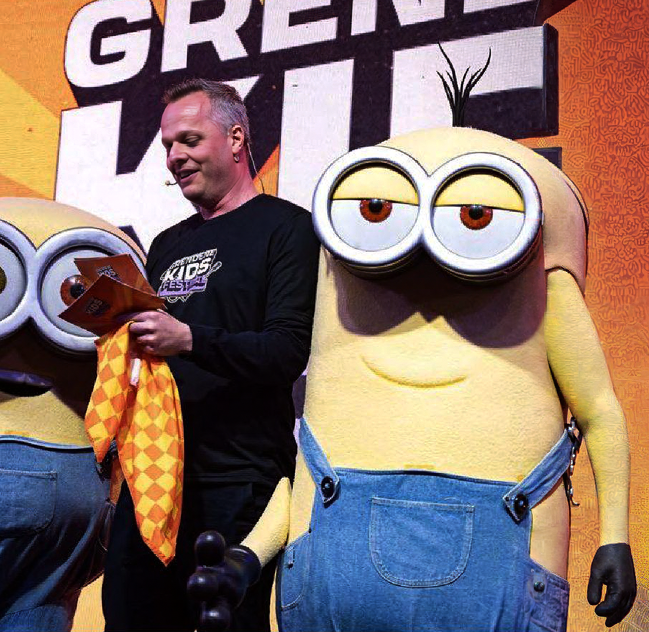 Homem com cabelo curto e pele clara sorrindo, vestindo uma camiseta preta, segurando um troféu dourado e uma folha de papel. Ao fundo, um mural com o logo do Minions e uma frase parcialmente visível, com fundo laranja e detalhes decorativos.