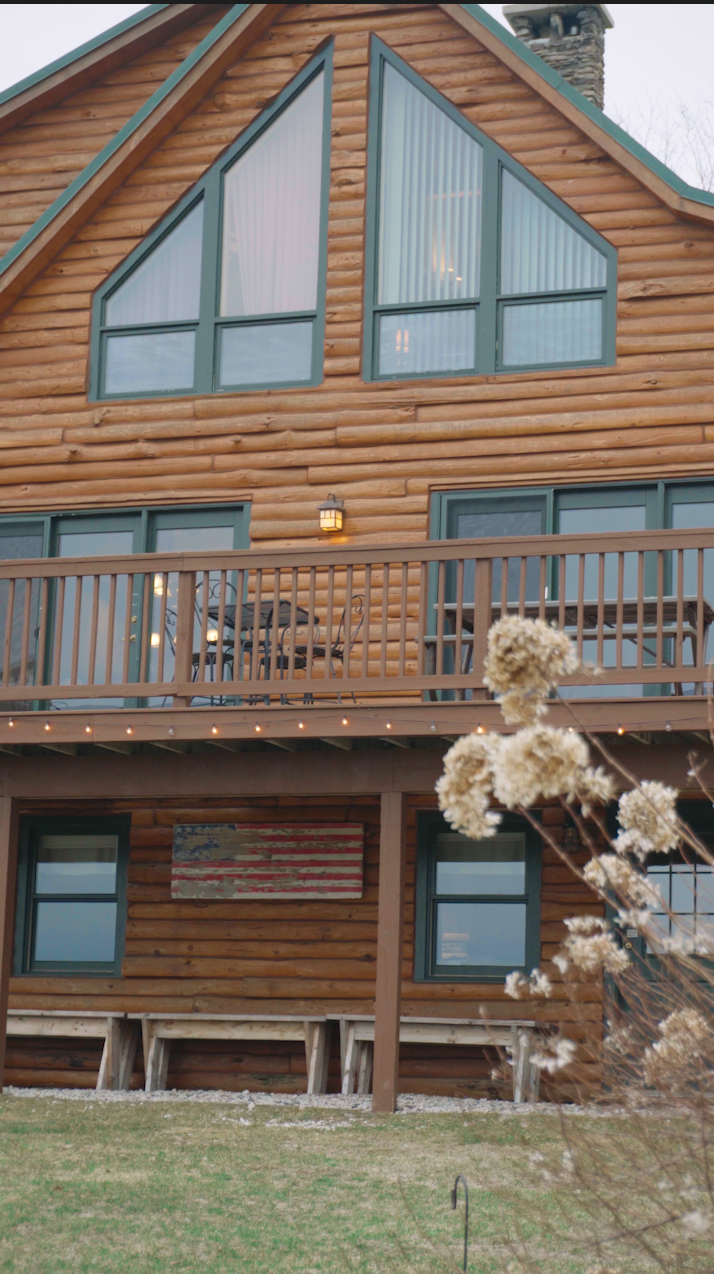 Wooden house with large angled windows in the upper floor, a balcony with outdoor furniture, an American flag decoration on the wall, and drying benches beneath. There are dried flowers in the foreground.