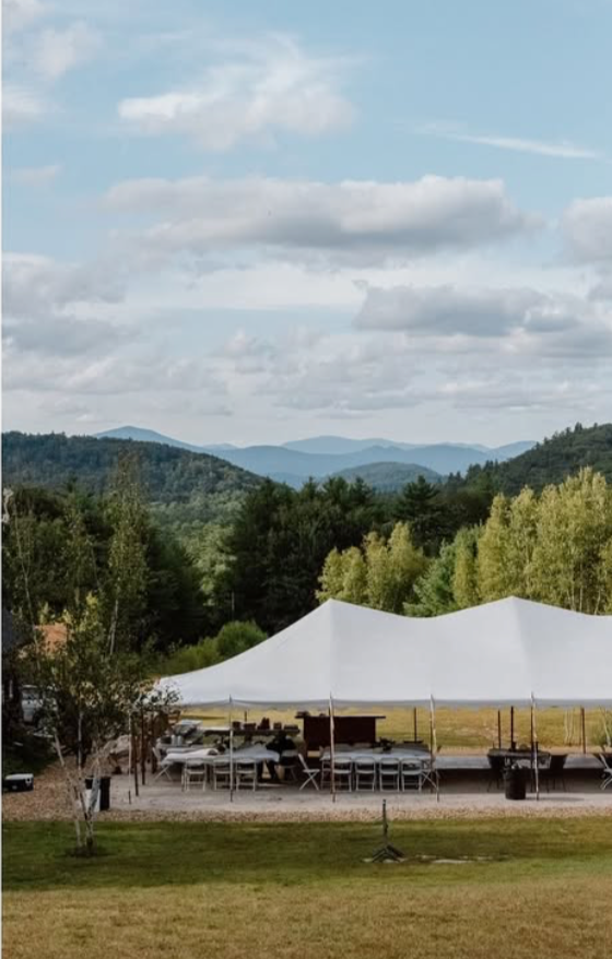 An outdoor event setup with a large white tent, tables, and chairs on a grassy area with trees and mountains in the background under a partly cloudy sky.