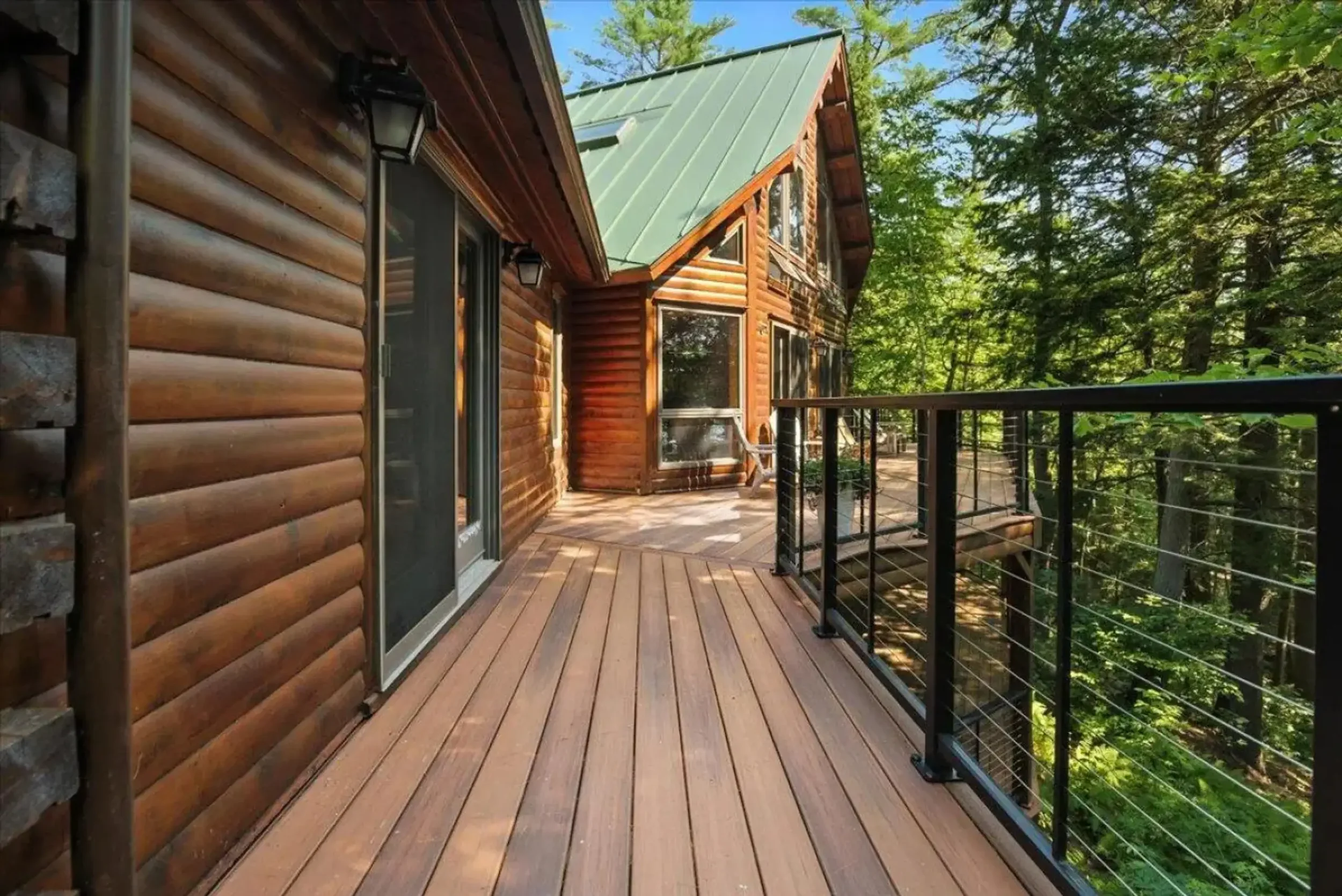 Wooden deck attached to a log cabin house with glass sliding door, black metal railing, surrounded by green trees, under a blue sky.
