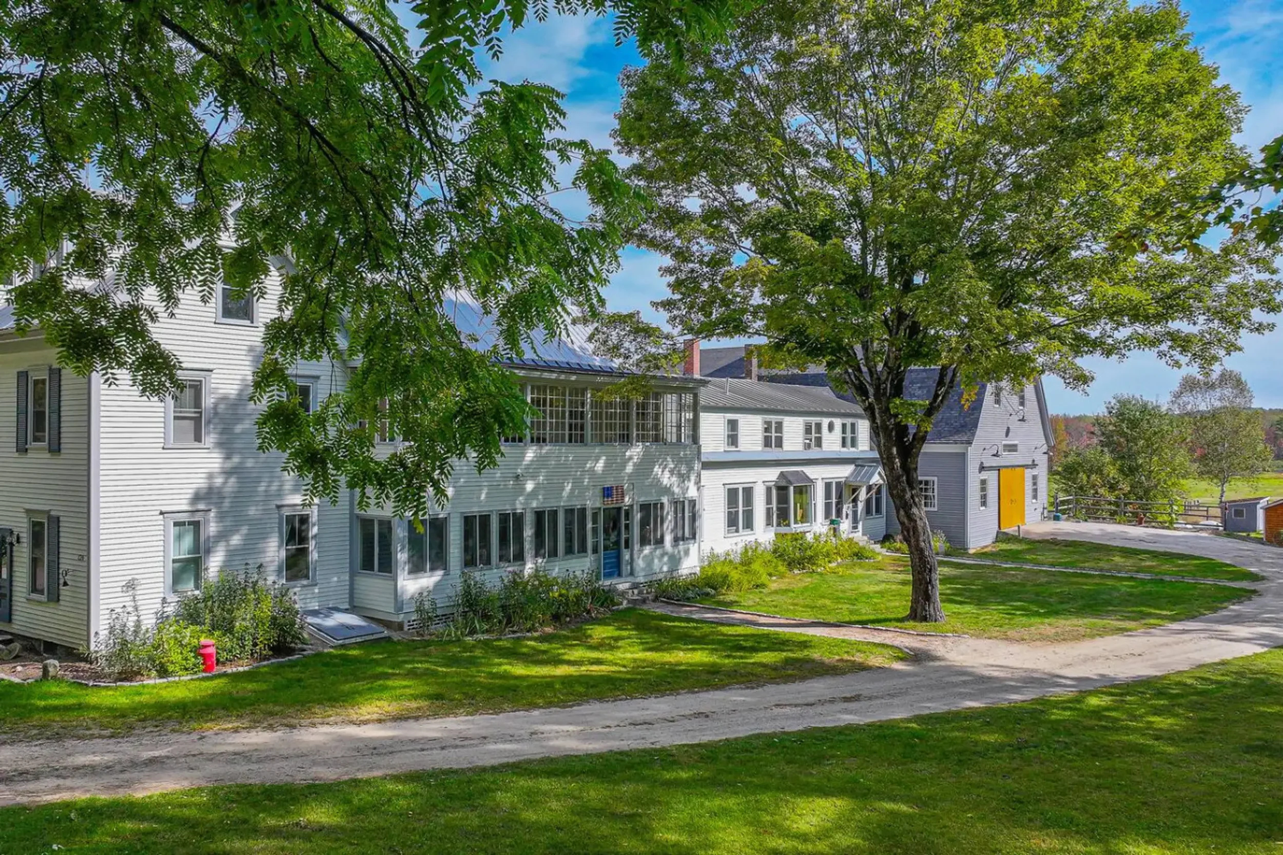A residential area with white houses featuring numerous windows, surrounded by green trees and lawns, under a partly cloudy sky.