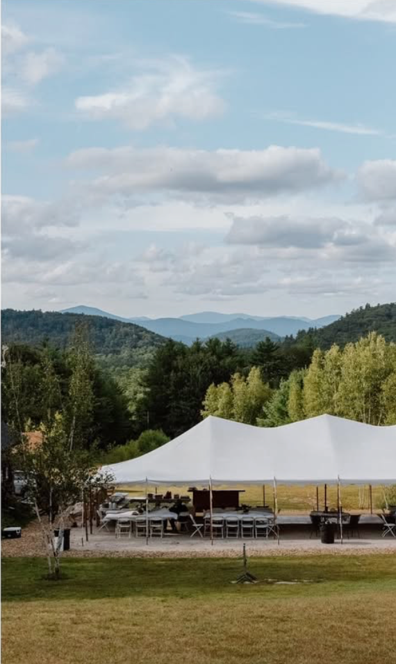 A large white event tent set up outdoors on a grassy area, with chairs and tables underneath, surrounded by trees and a scenic mountain landscape in the background.