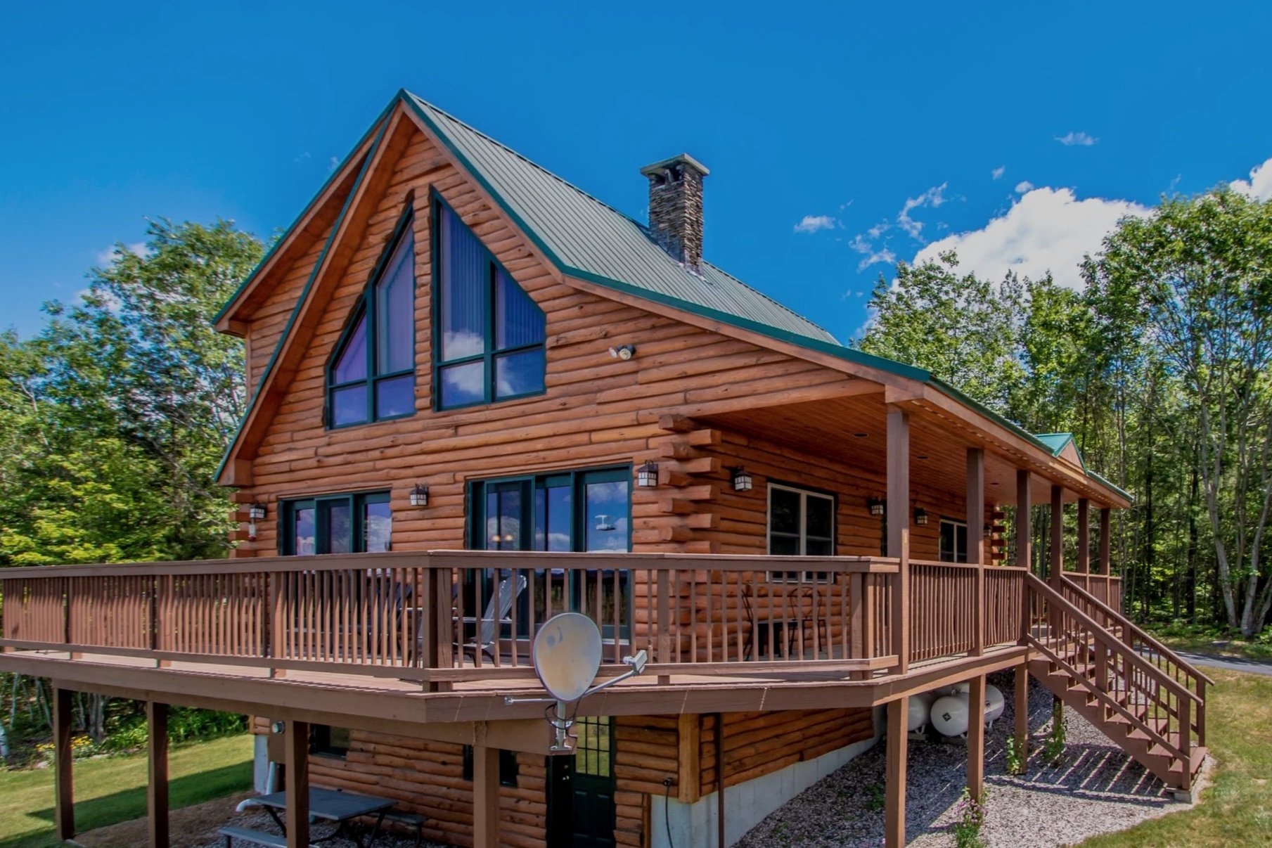 A two-story wooden house with a deck, large windows, and a metal roof, surrounded by trees, under a blue sky with some clouds.