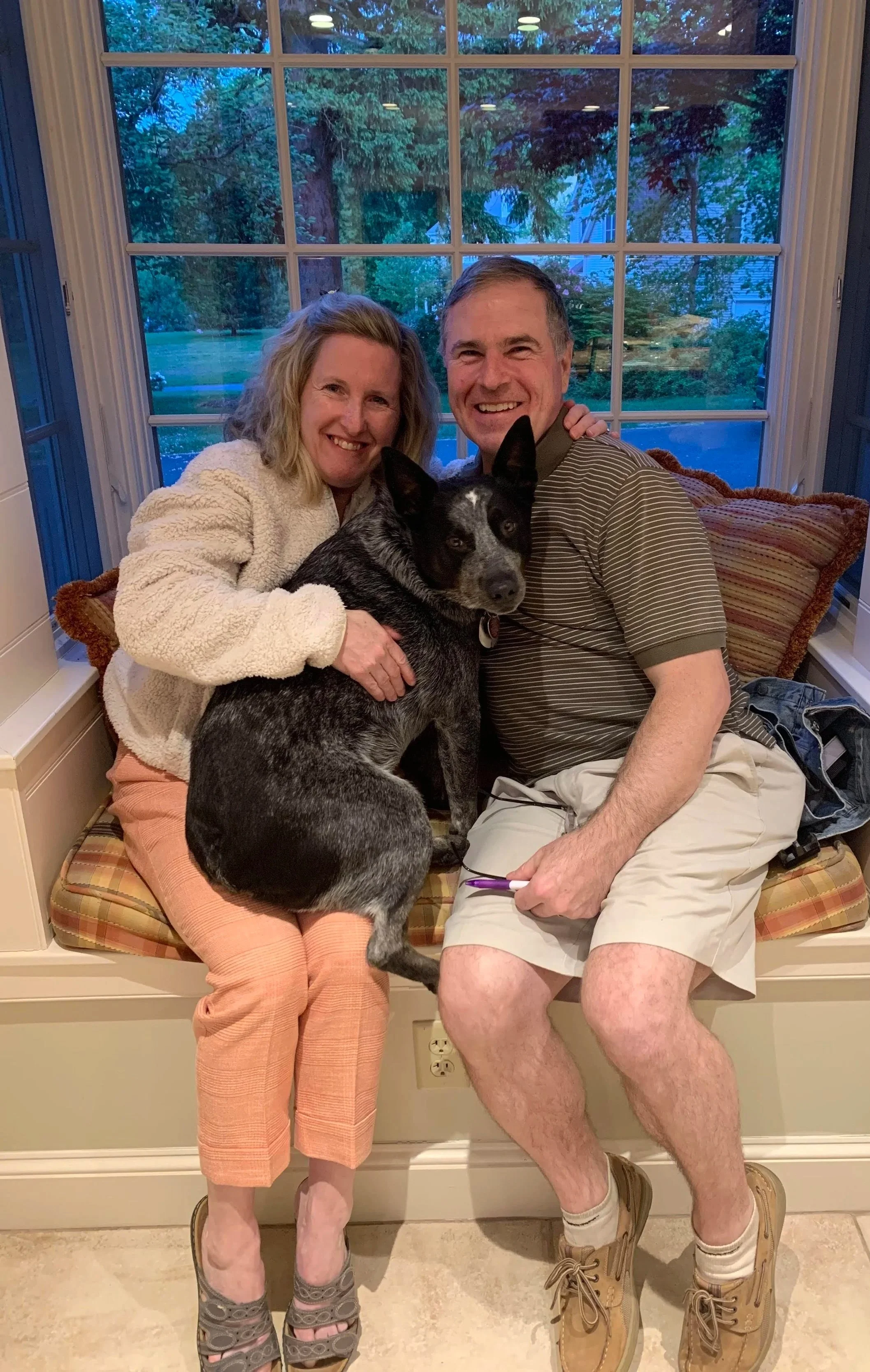 Smiling couple sitting on a window seat with their dog. The woman is wearing a cream fuzzy jacket and peach pants, and the man is wearing a striped polo shirt and beige shorts. The dog is a black and grey, medium-sized, short-haired breed with a white patch on its face. The background shows large window with trees outside.