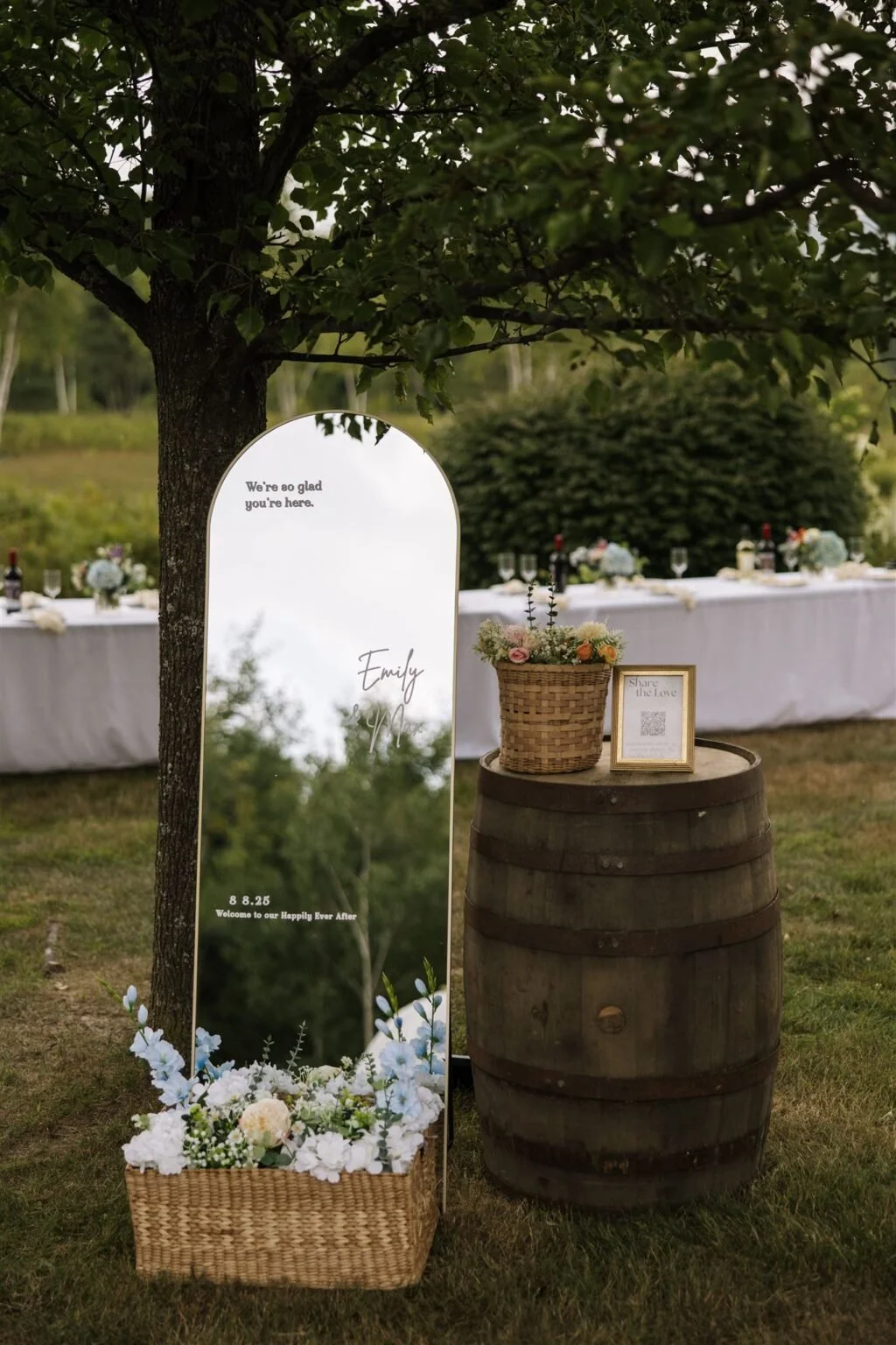 Outdoor wedding reception setup with a tree and a tall mirror sign that reads, 'We're so glad you're here,' with the names Emily and an unreadable last name, and the date 8.8.25. A wicker basket with white and pastel flowers is at the base of the mirror, and a small frame with a QR code is on a wooden barrel next to a flower arrangement in a wicker basket. In the background, a long white table is set with floral centerpieces, wine bottles, and glassware.