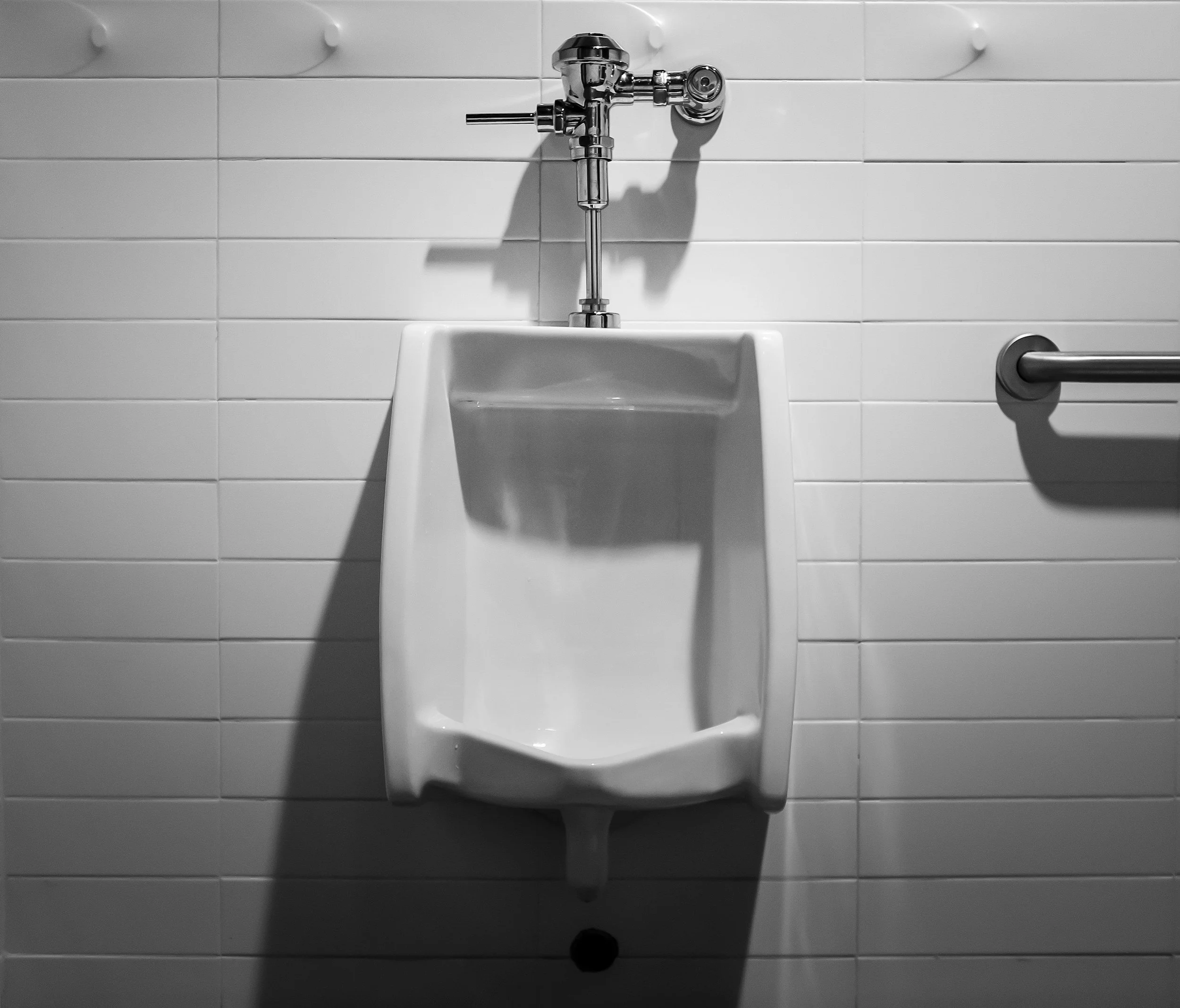A white urinal with a chrome flush valve mounted on a white tiled wall.