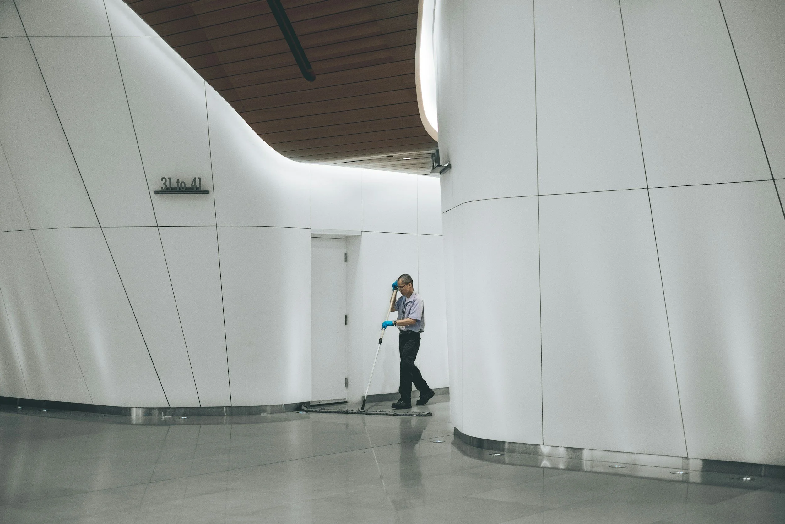 A man wearing glasses, a gray shirt, and black pants cleaning the polished floor of a modern building with white walls and wooden ceiling panels. He is using a mop and wearing blue gloves.