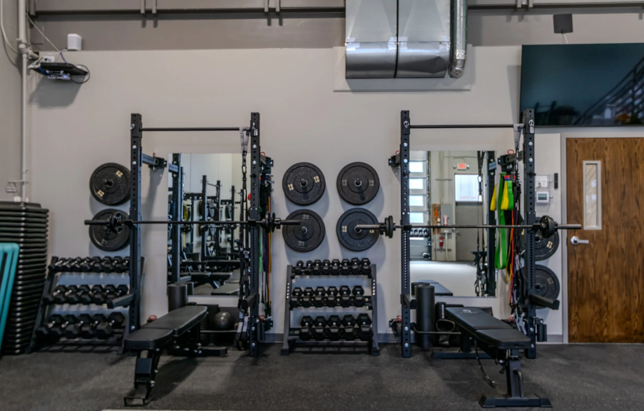 Interior of a gym with weightlifting racks, weights, and colorful resistance bands. Mirrors line the wall, and workout equipment is visible, including dumbbells and a bench.