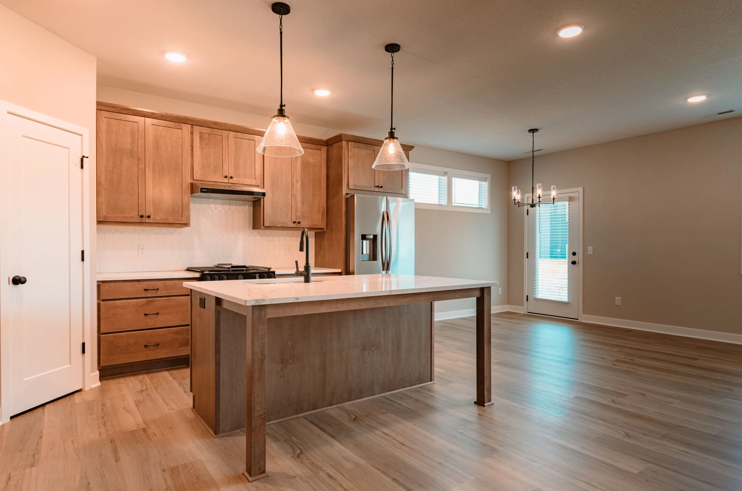 Open-concept kitchen with wooden cabinets, an island with a marble top, pendant lights, and a door leading outside, with hardwood floors and neutral wall color.