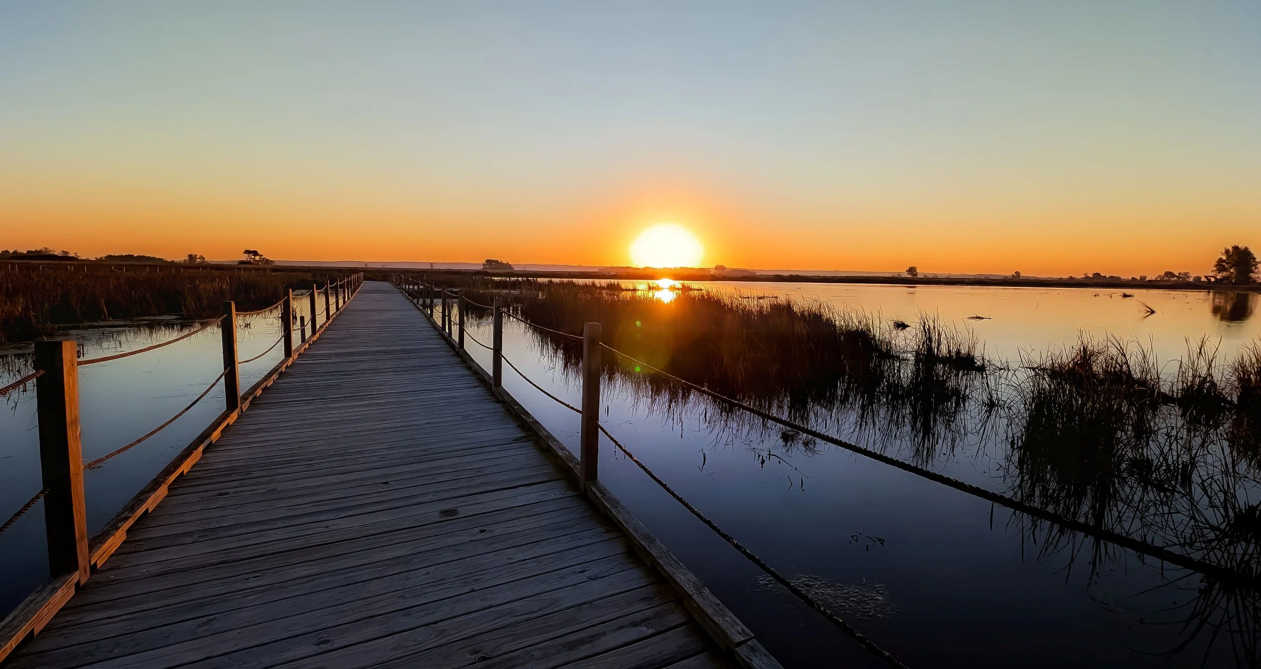 A wooden boardwalk over a waterway at sunset, with calm water reflecting the orange and yellow sky and distant trees on the horizon.