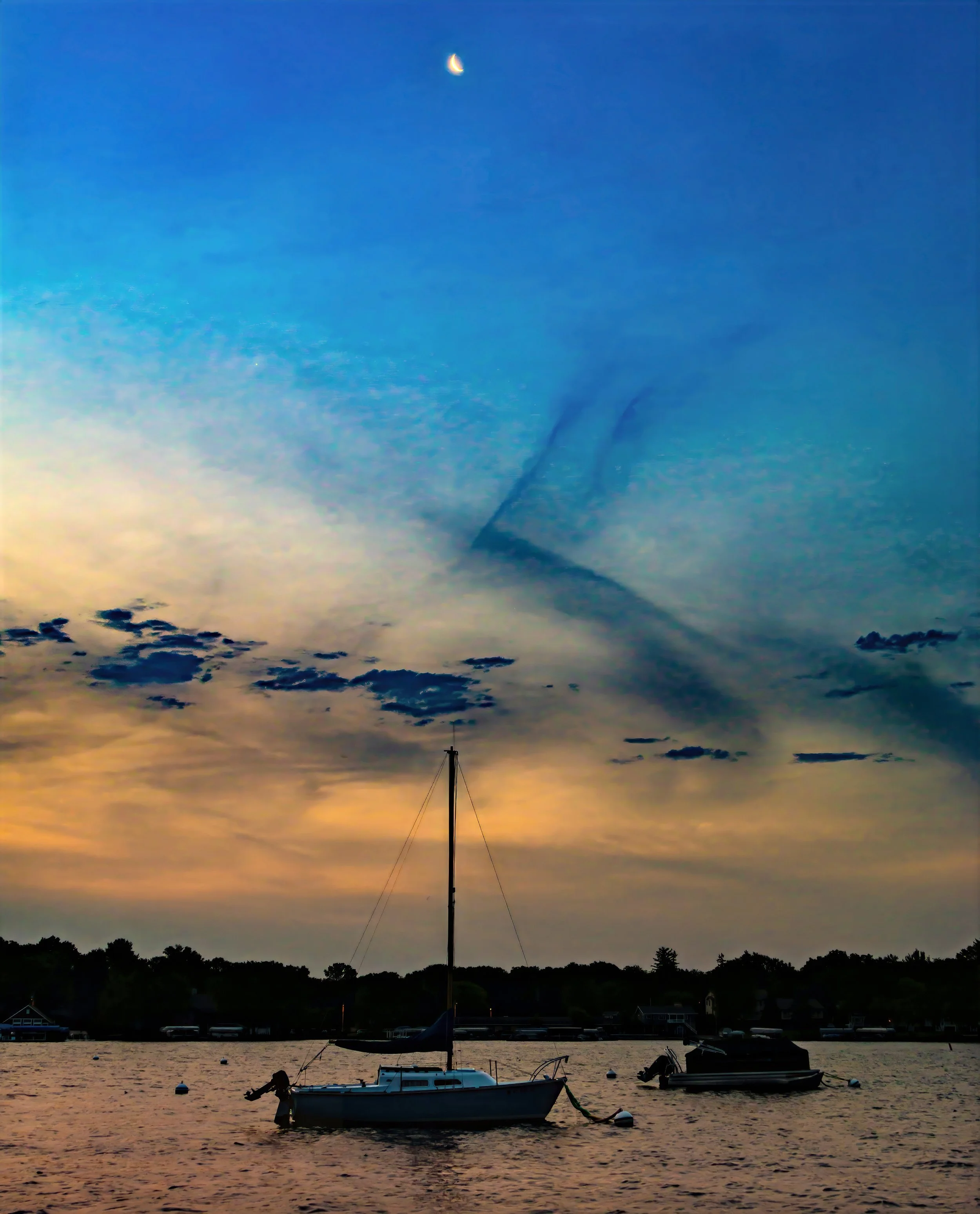 Boats anchored in a calm body of water during sunset, with trees and houses in the background, and a colorful sky with clouds and a crescent moon.