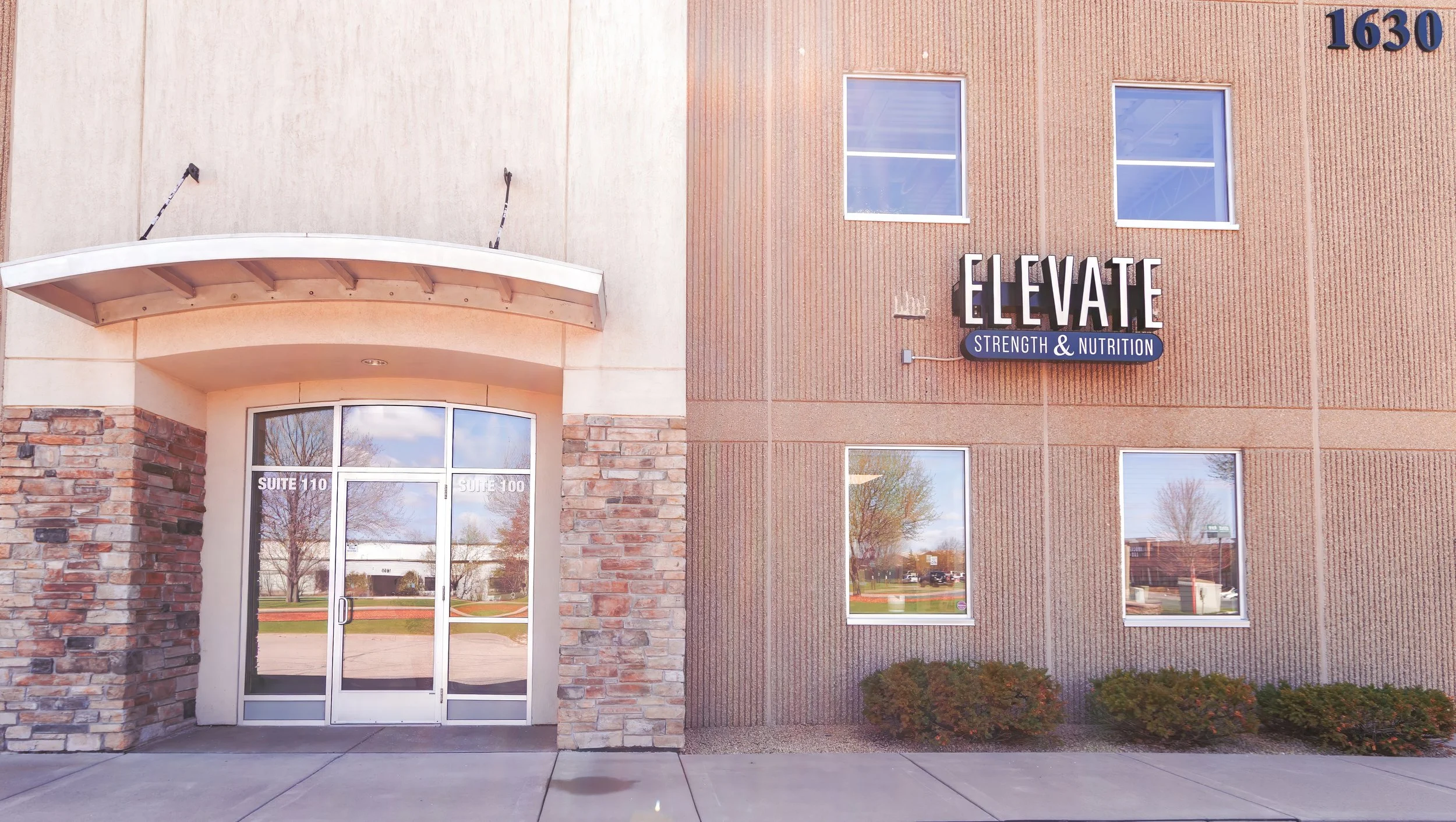 Exterior of a commercial building with a brick facade, large windows, and a sign that reads 'ELEVATE Strength & Nutrition' near the top right corner. The entrance has glass doors with the suite numbers 'Suite 110' and 'Suite 100' on the door frames. 