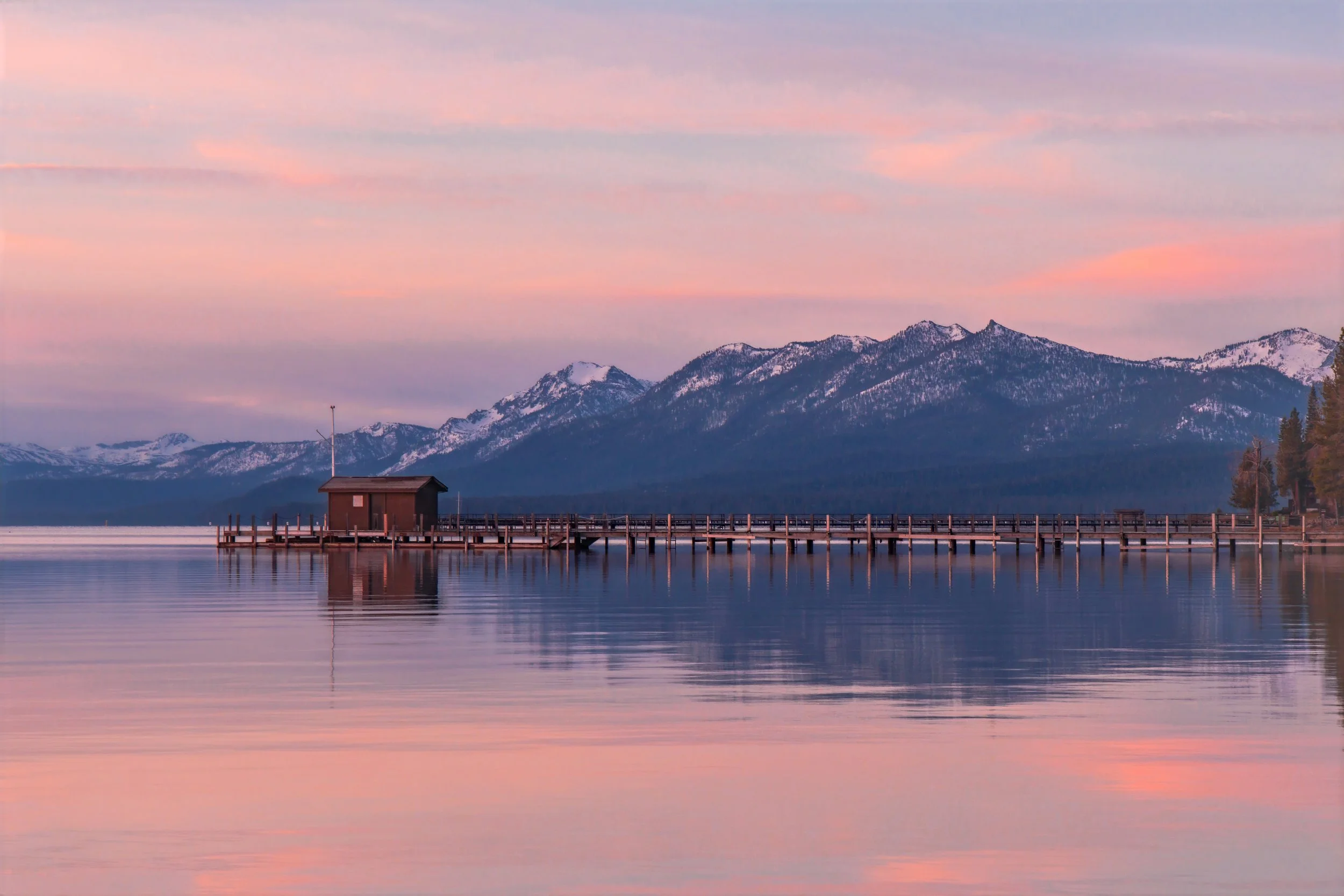 A calm lake reflecting a pastel-colored sky at sunset with mountains in the background, a small wooden structure on a pier extending into the water, and trees along the shoreline.