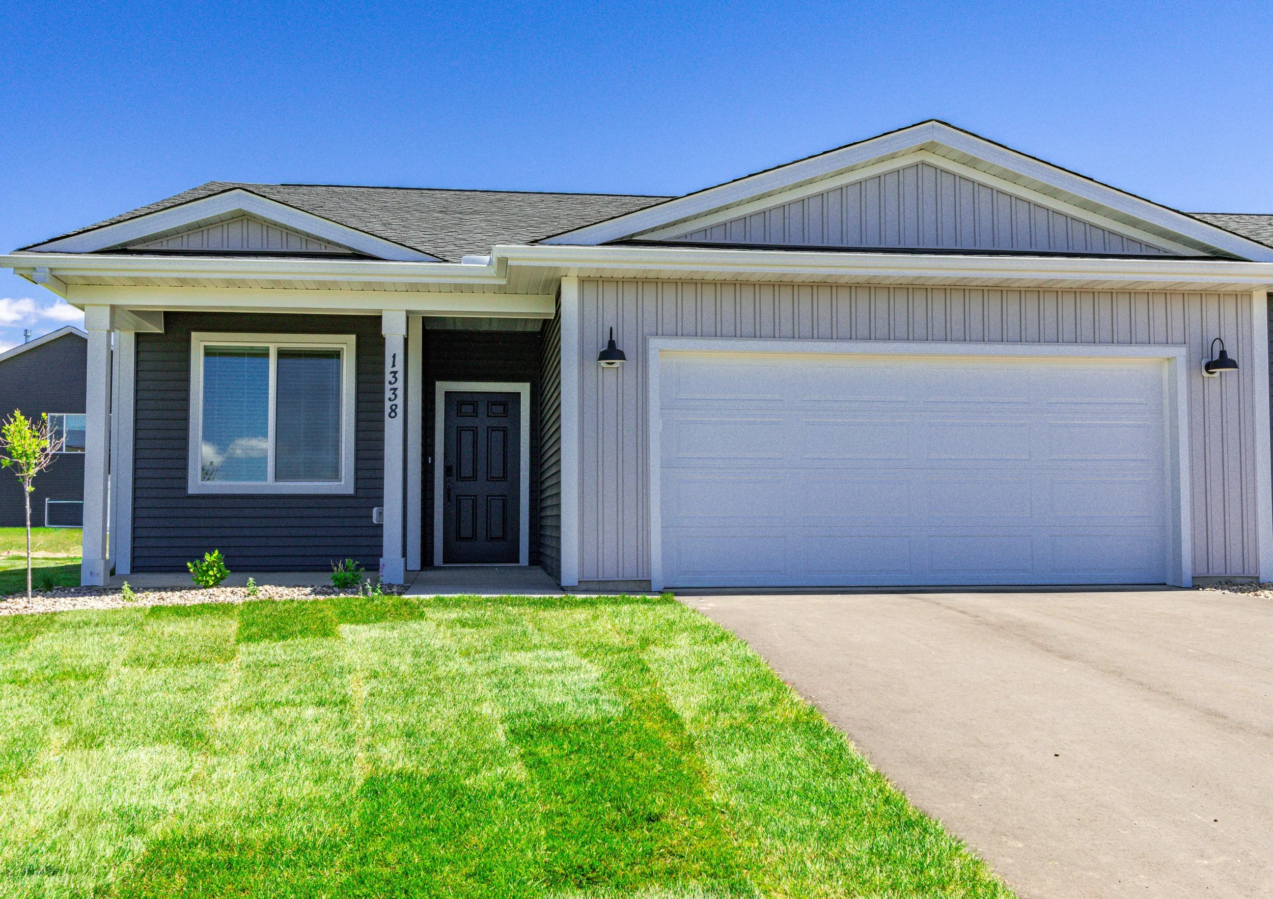 Front view of a modern house with gray and white siding, a black front door, large front window, attached garage, and a well-maintained green lawn under a clear blue sky.