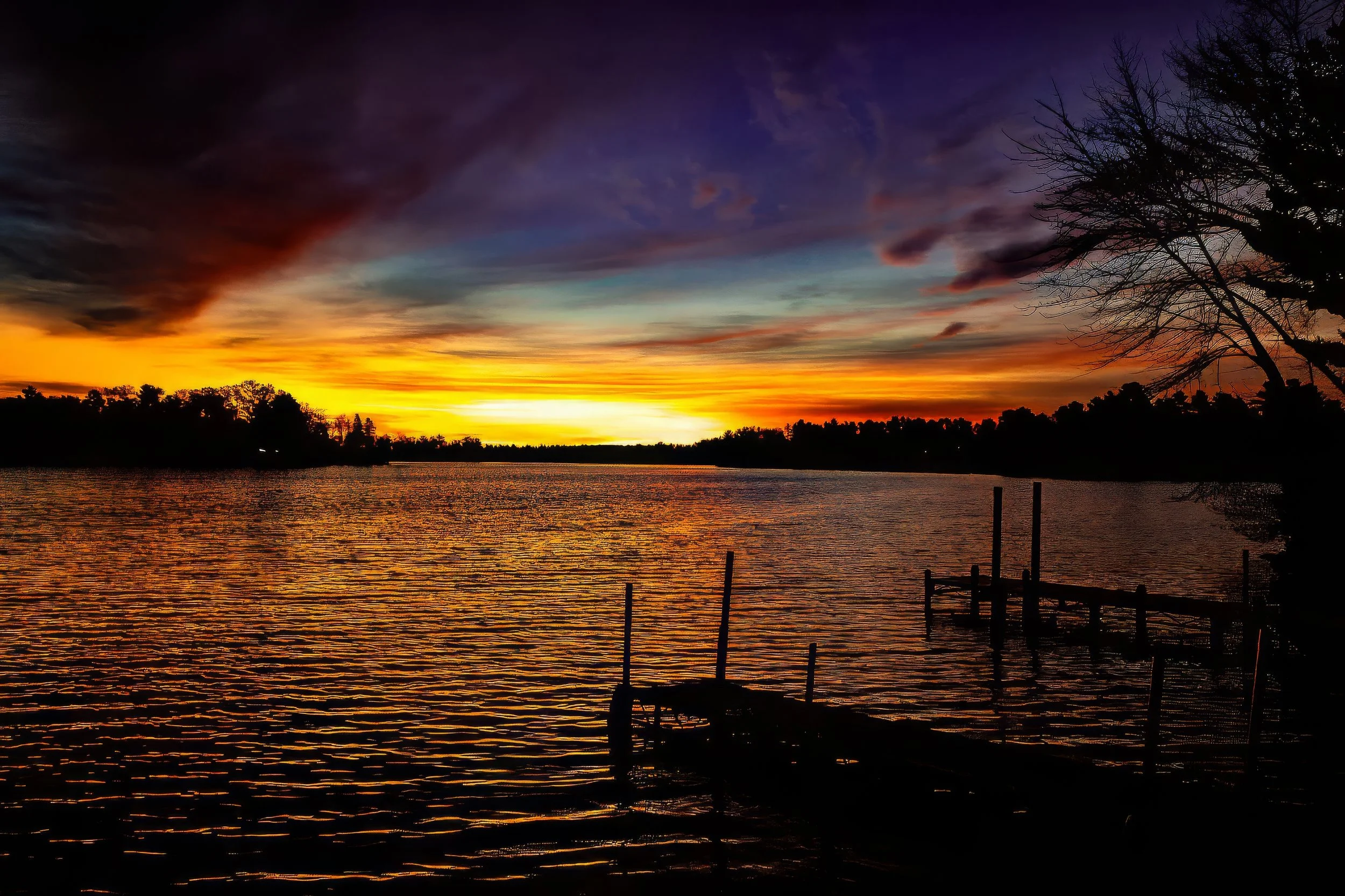 Sunset over a river with orange, purple, and blue sky, silhouetted trees, and a small dock or pier in the foreground.