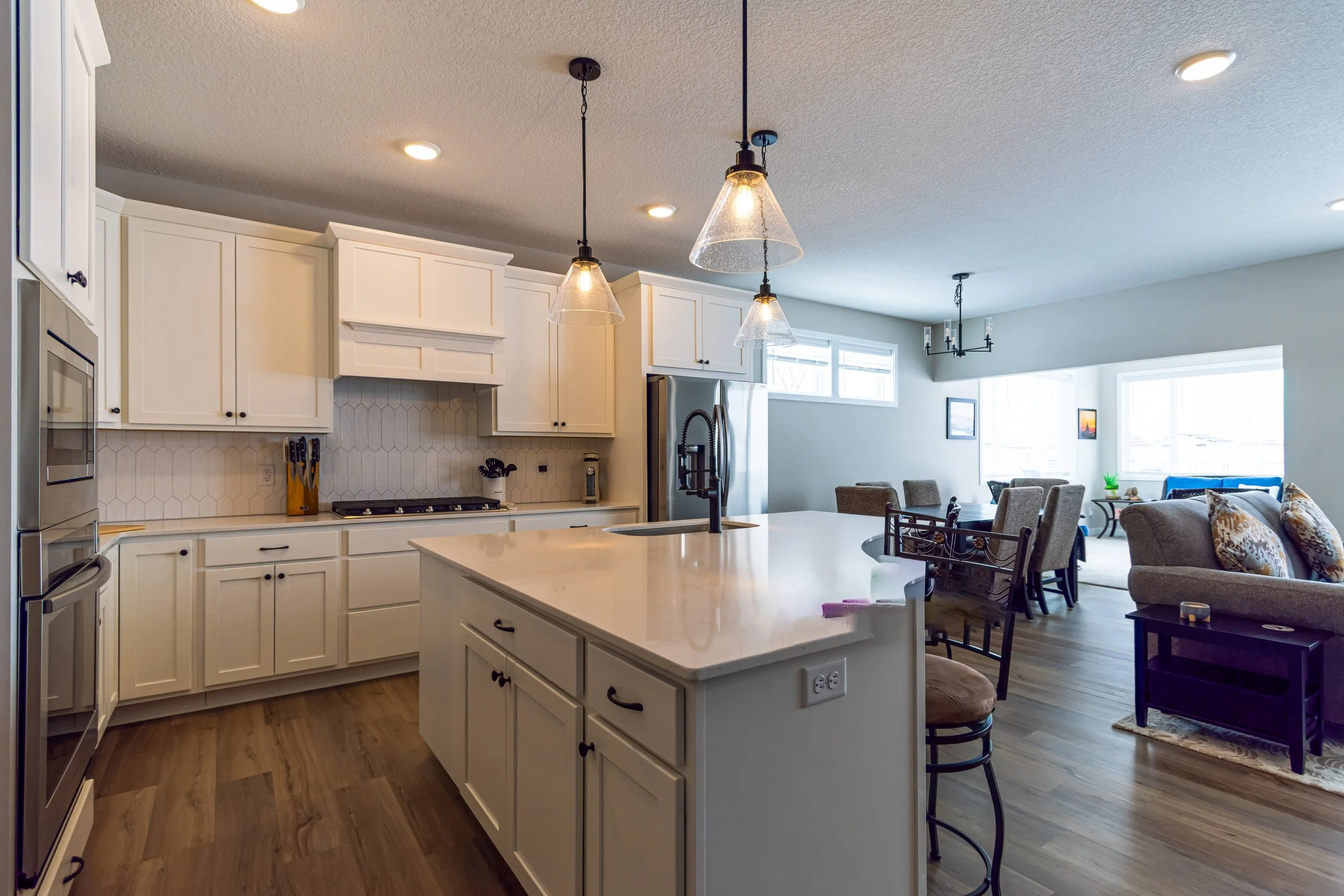 Open kitchen with white cabinets, a kitchen island, stainless steel refrigerator, and modern hanging pendant lights. The adjacent living room has large windows, a chandelier, and comfortable seating.