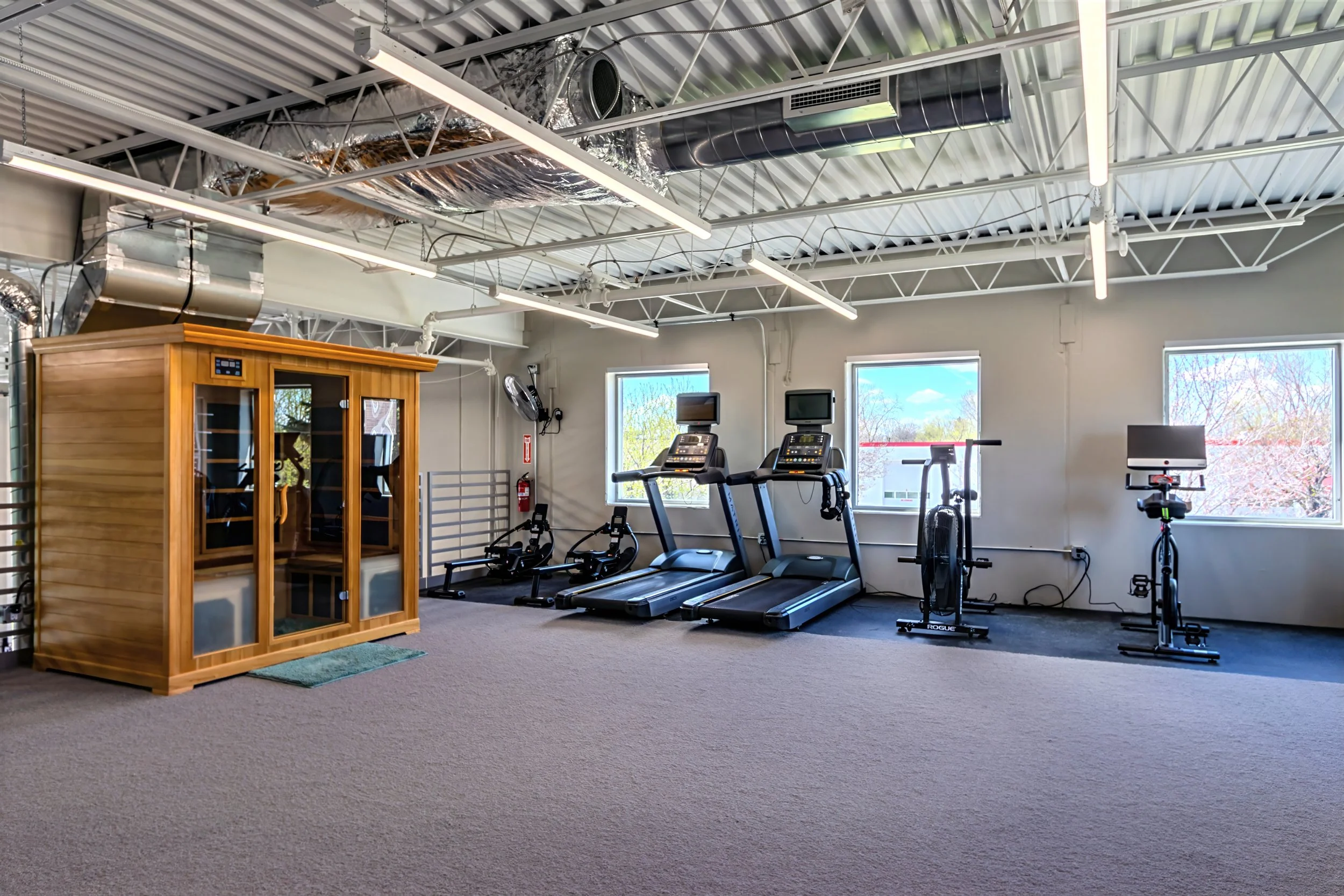 A gym with treadmill and workout equipment near windows, with an infrared sauna in the foreground, and ceiling with exposed ductwork and lighting.