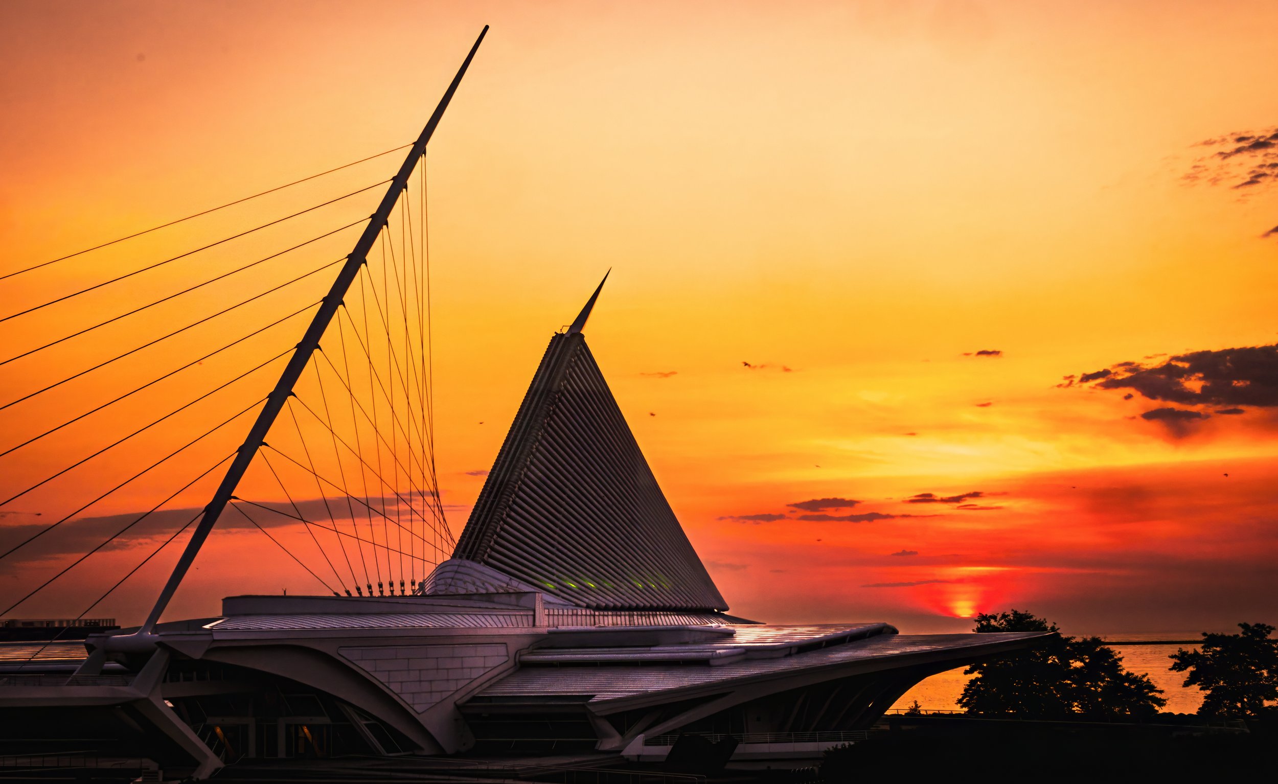 Modern architectural building with sail-like roof structure and tall spires against a vibrant orange sunset sky.
