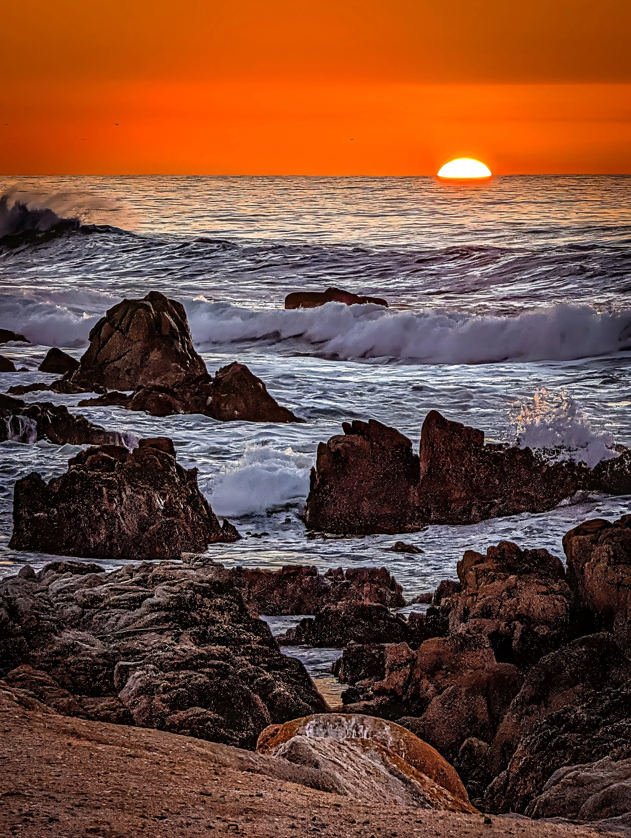A sunset over the ocean with rocks in the foreground and a vibrant orange sky.