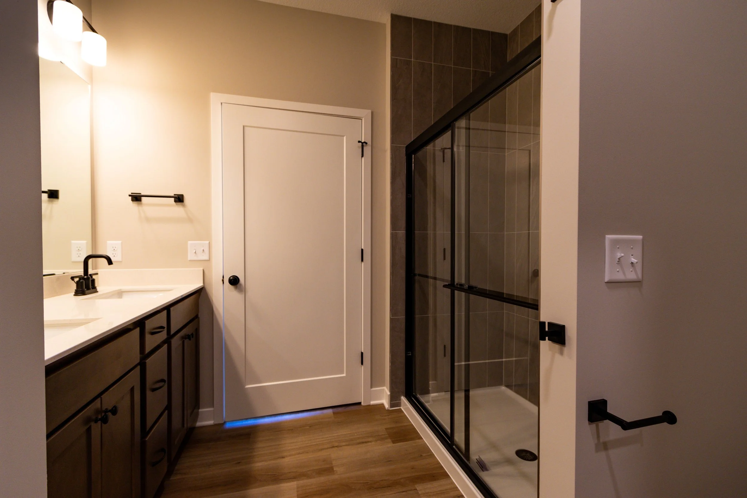 A bathroom vanity with dark wooden cabinets, a white countertop, and a black faucet. Next to it is a white door and a glass-enclosed shower with brown tile. The wall has a towel ring and electrical outlets.