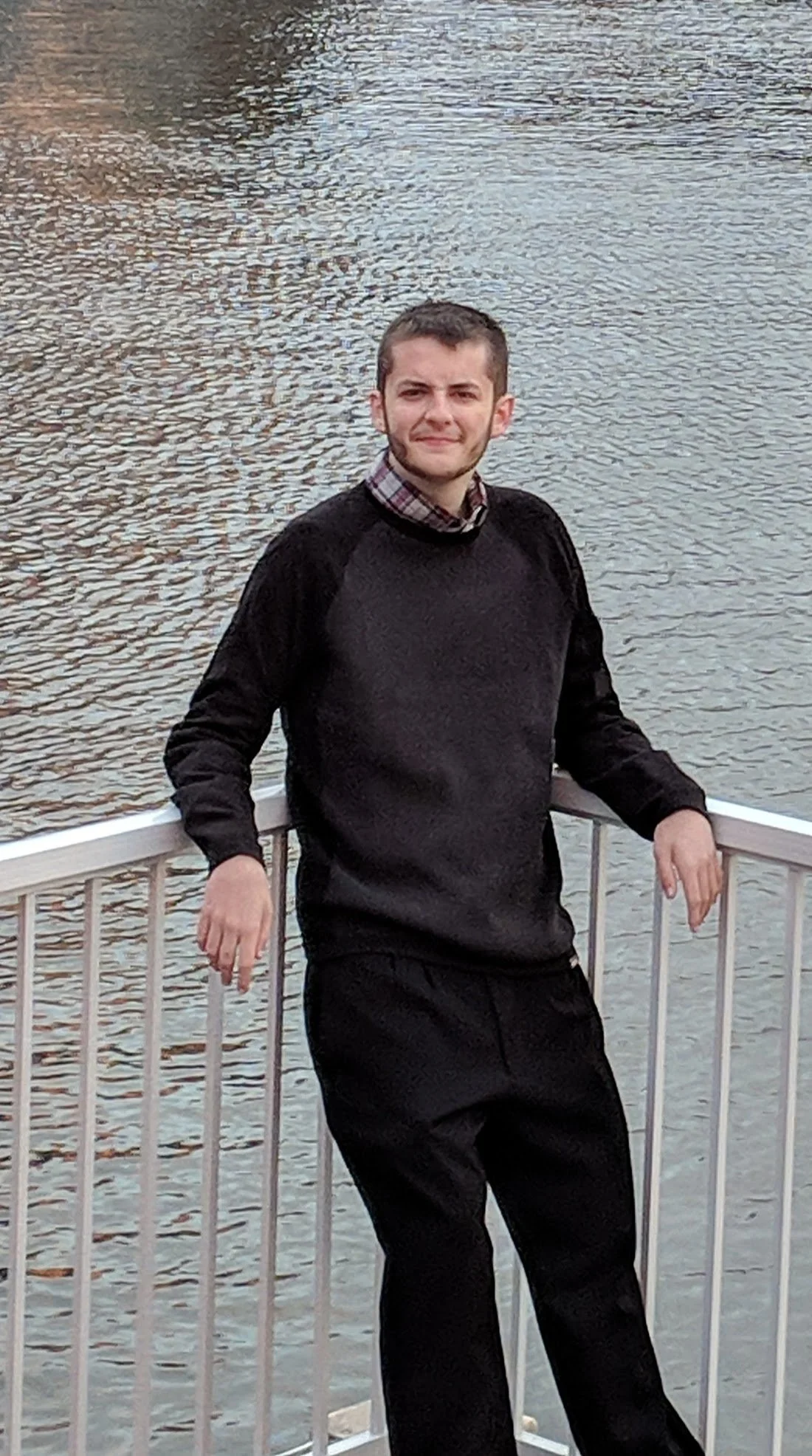 A young man with short dark hair and a beard standing on a boat railing next to a body of water during daytime, wearing a black sweater and black pants.