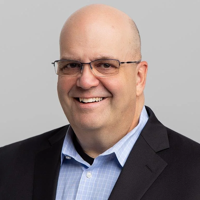 Headshot of a smiling man wearing glasses, a black blazer, and a blue collared shirt against a plain gray background.