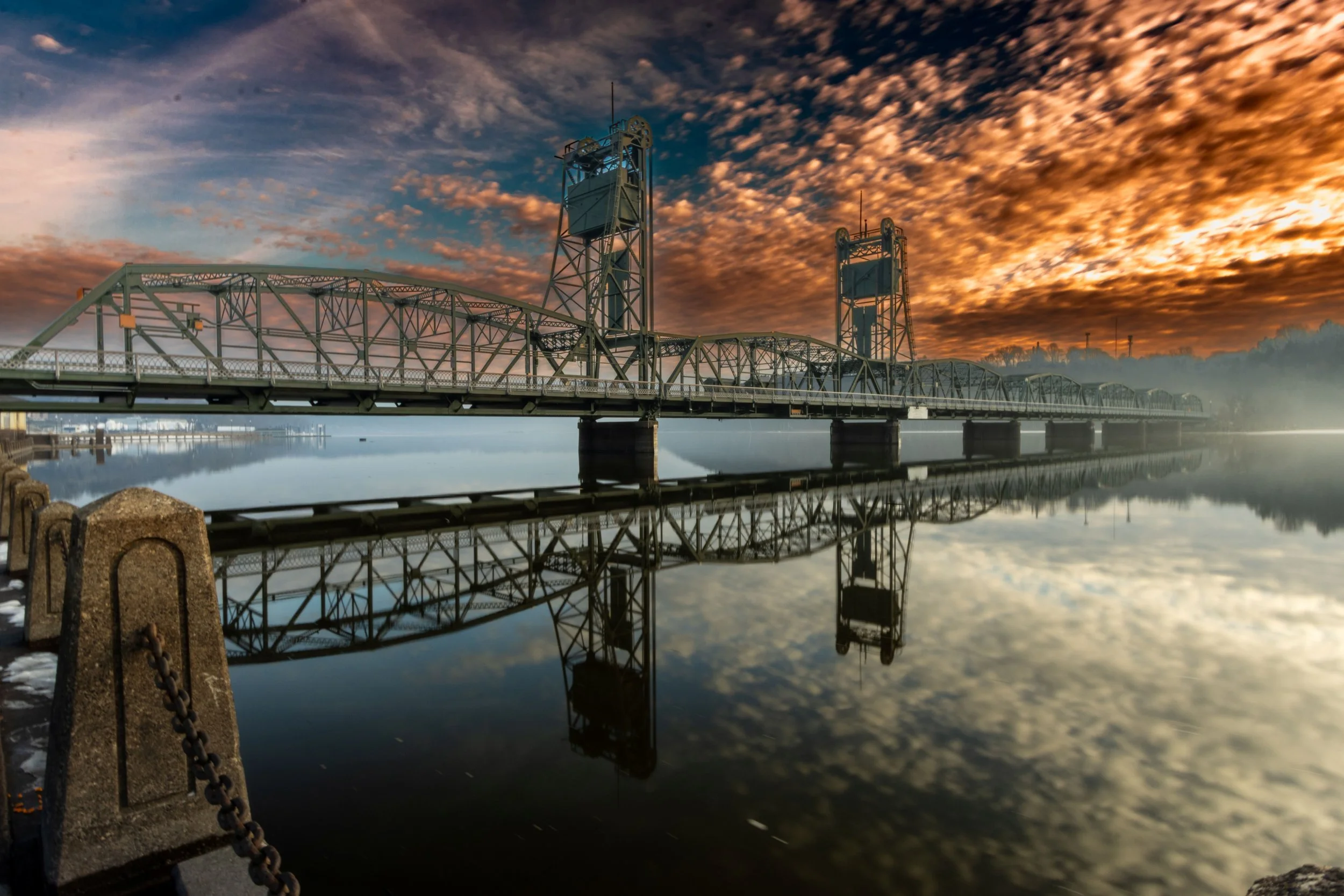 A bridge over calm water reflects the colorful red and orange sunset sky, with cloud formations.