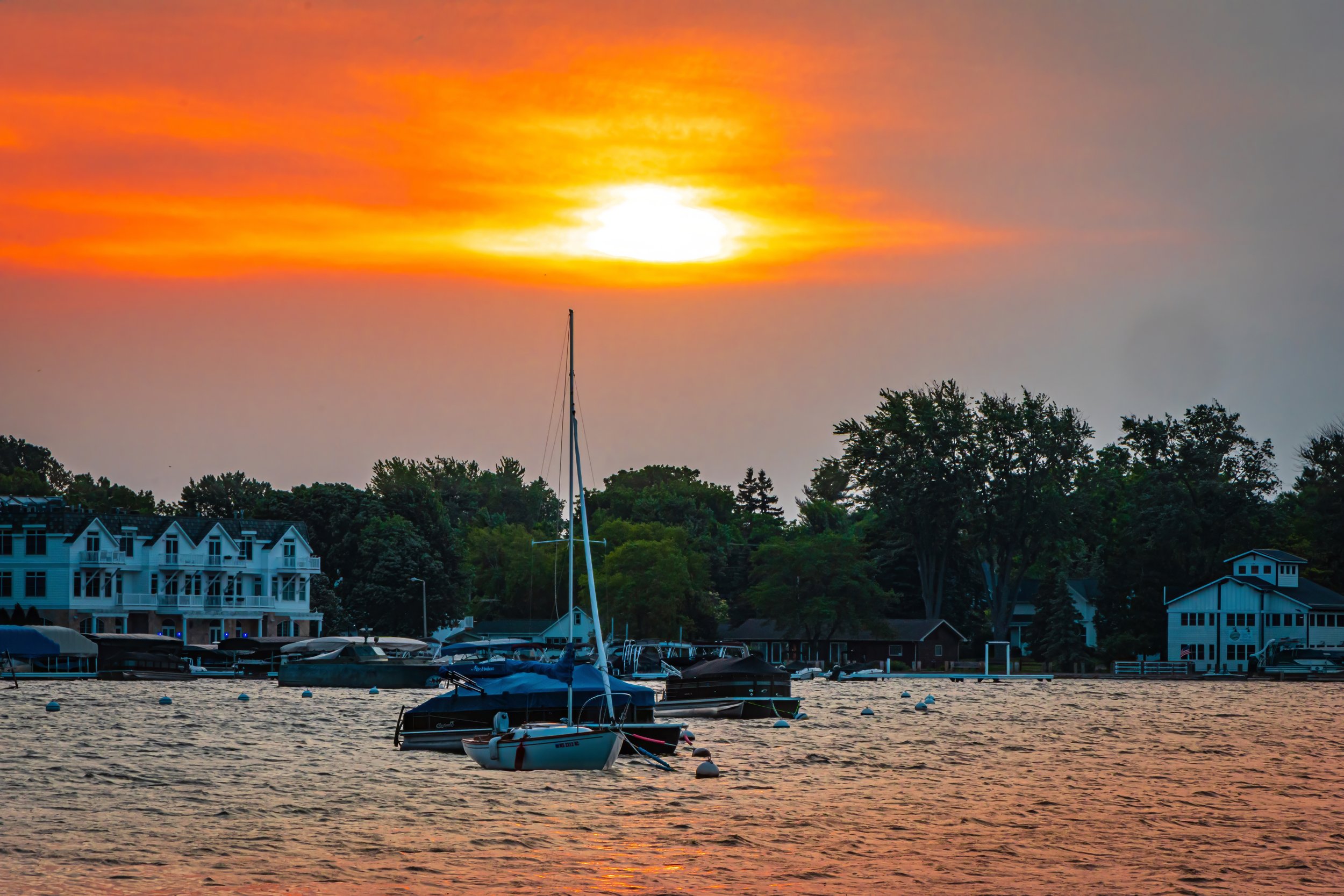 A marina at sunset with several boats docked on the water, A sailboat in the foreground, multi-story residential buildings and green trees in the background, and a fiery orange and yellow sky with clouds overhead.