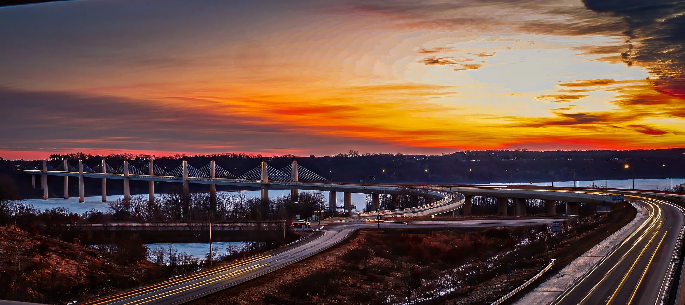 A highway bridge over a river at sunset with a colorful sky and light trails from moving vehicles.