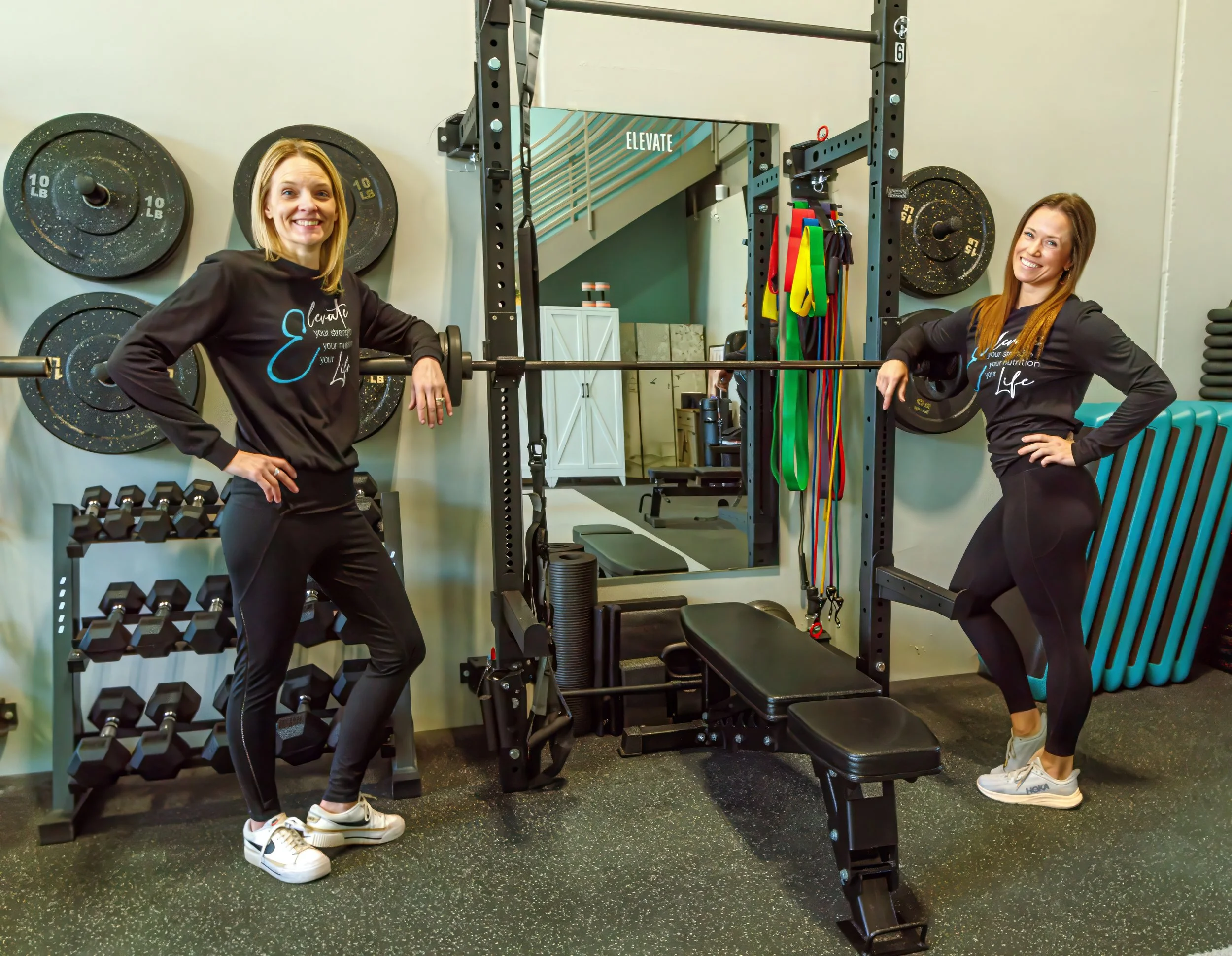 Two women in black workout clothes and white sneakers smiling in a gym near a squat rack with weight plates and colorful resistance bands.