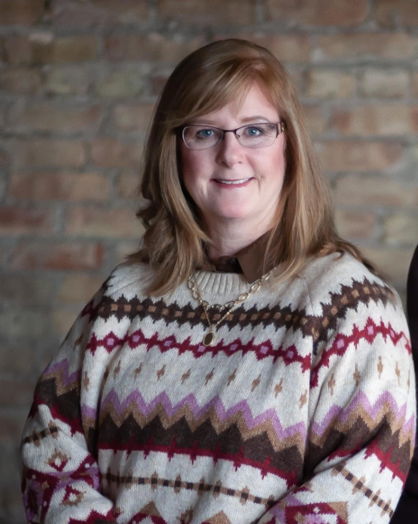 A woman with shoulder-length reddish-brown hair, wearing glasses, a patterned sweater, and a necklace, standing against a brick wall background.