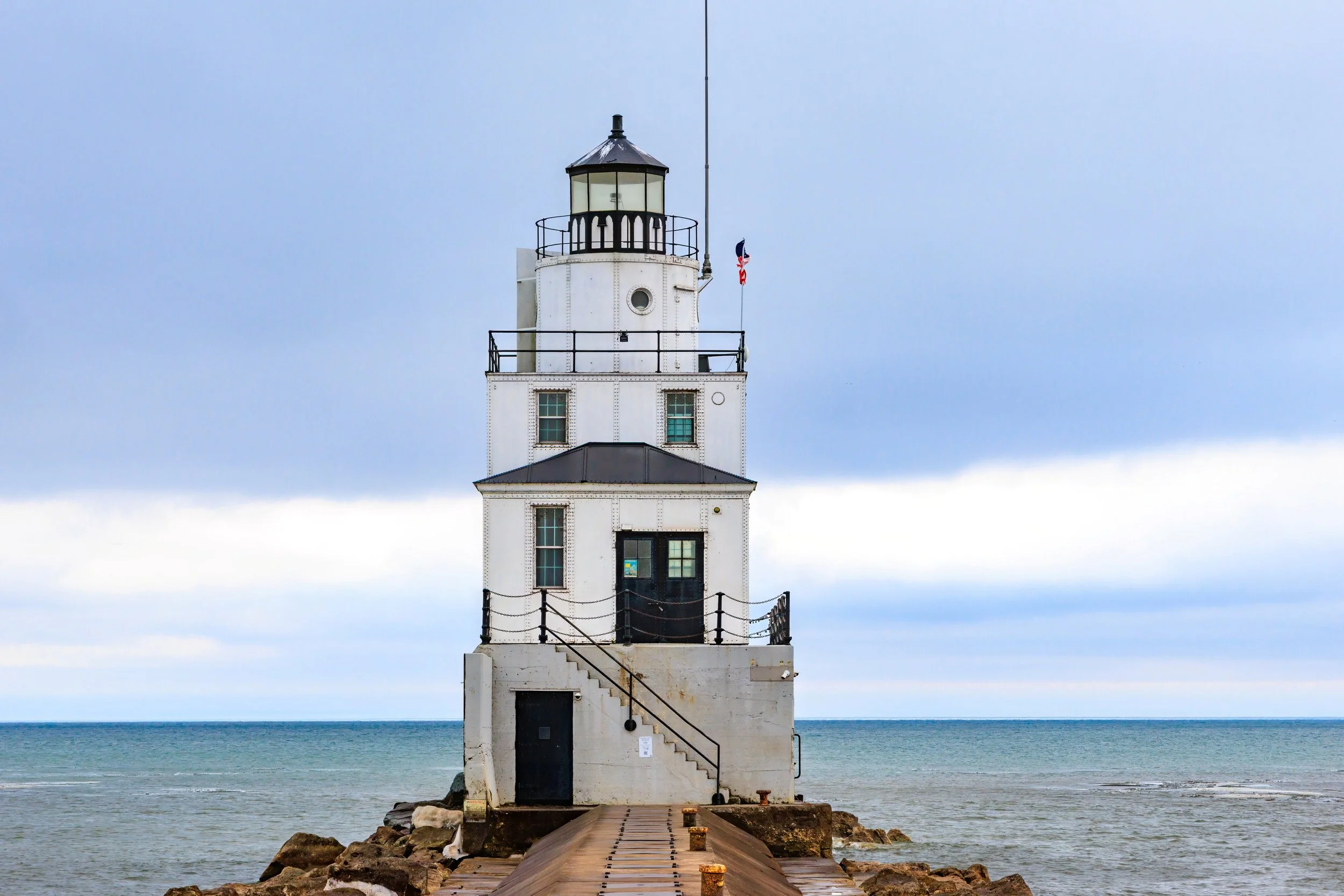 A white lighthouse at the end of a pier extending into the ocean, with a gray sky in the background.