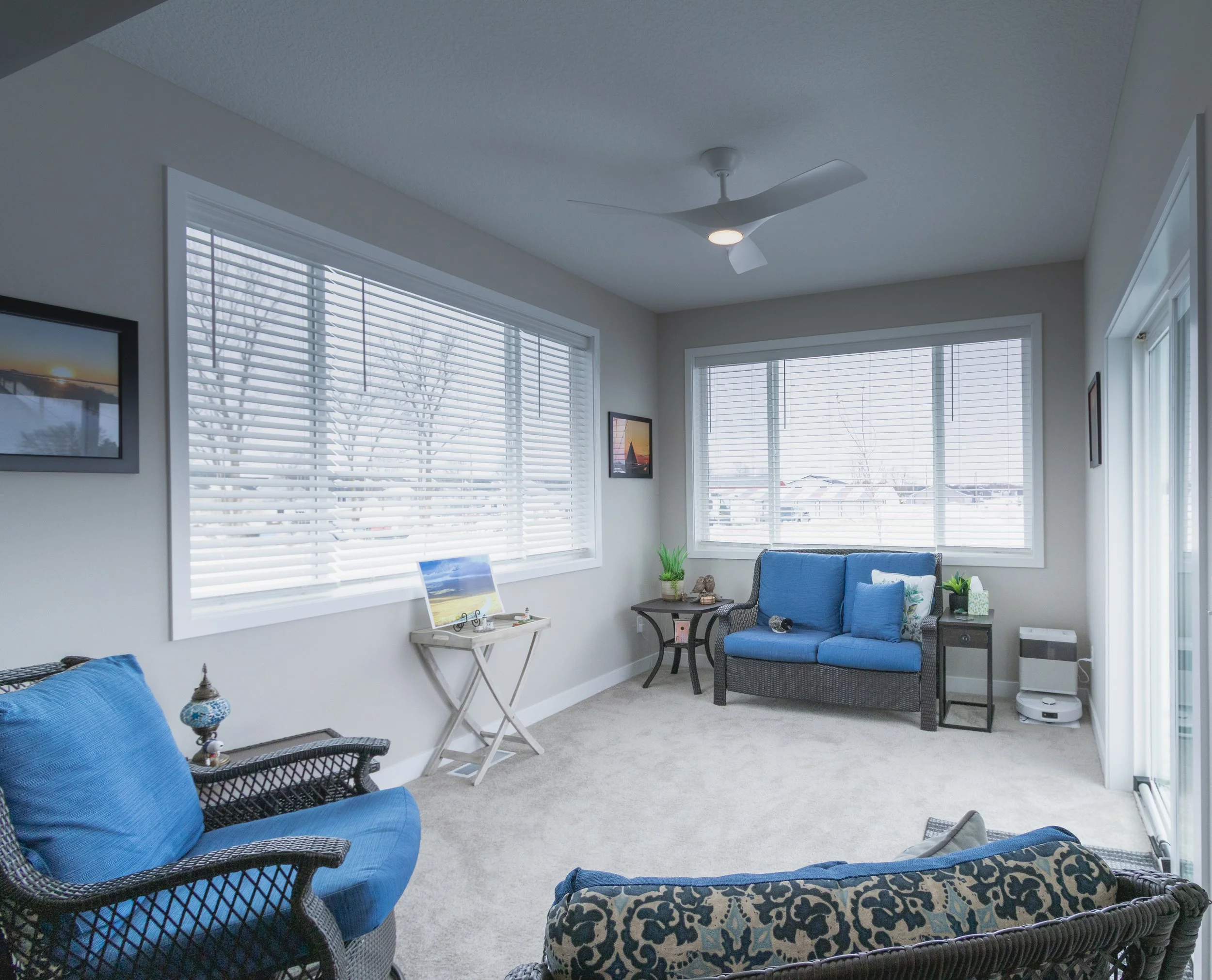 Bright living room with large windows, white blinds, a ceiling fan, framed pictures on the wall, blue sofas, a small side table, and decorative plants.