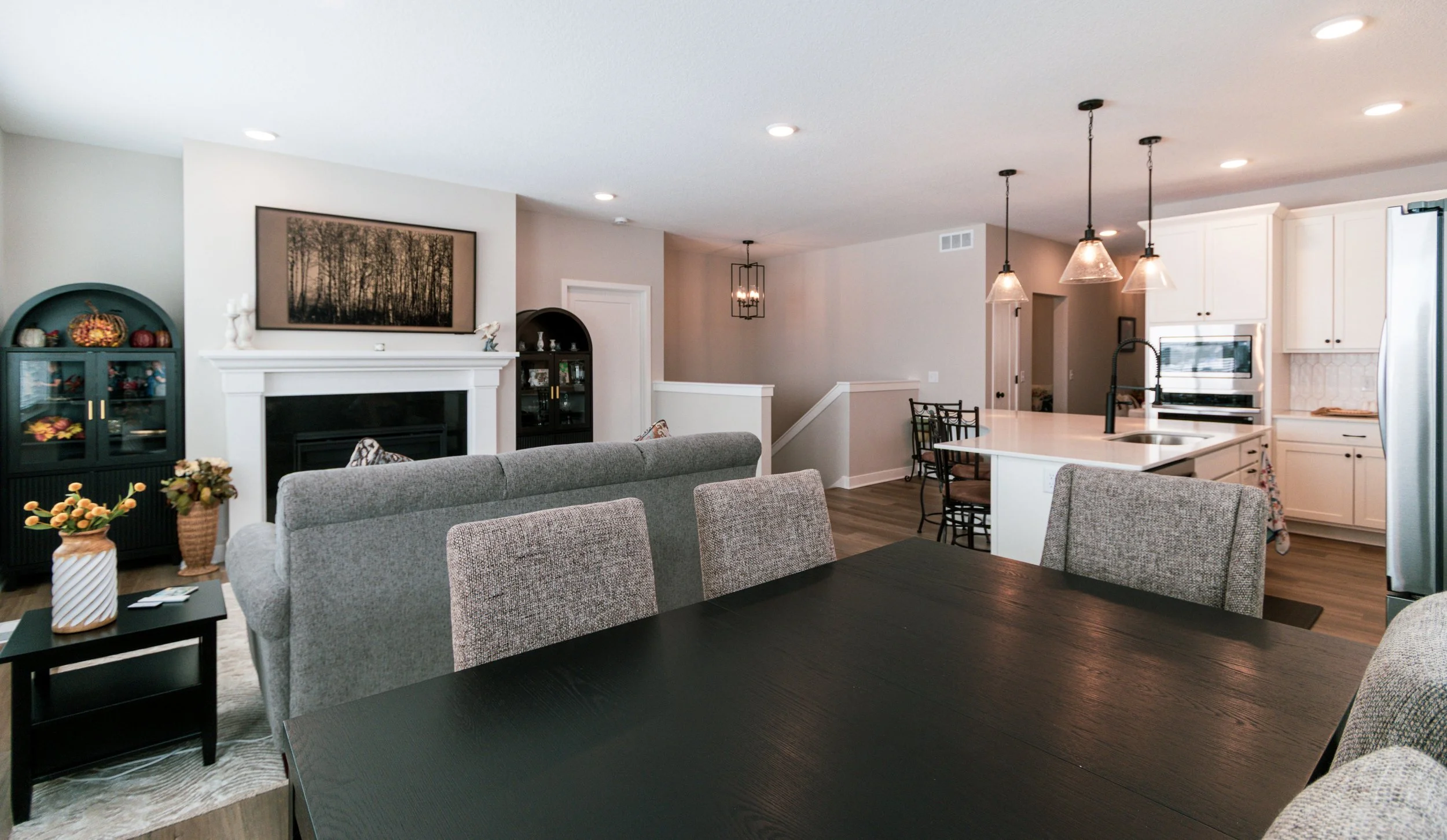 Open floor plan living area with gray sofas, black dining table with chairs, white kitchen island with hanging pendant lights, fireplace with decor, and wall art in a modern style.