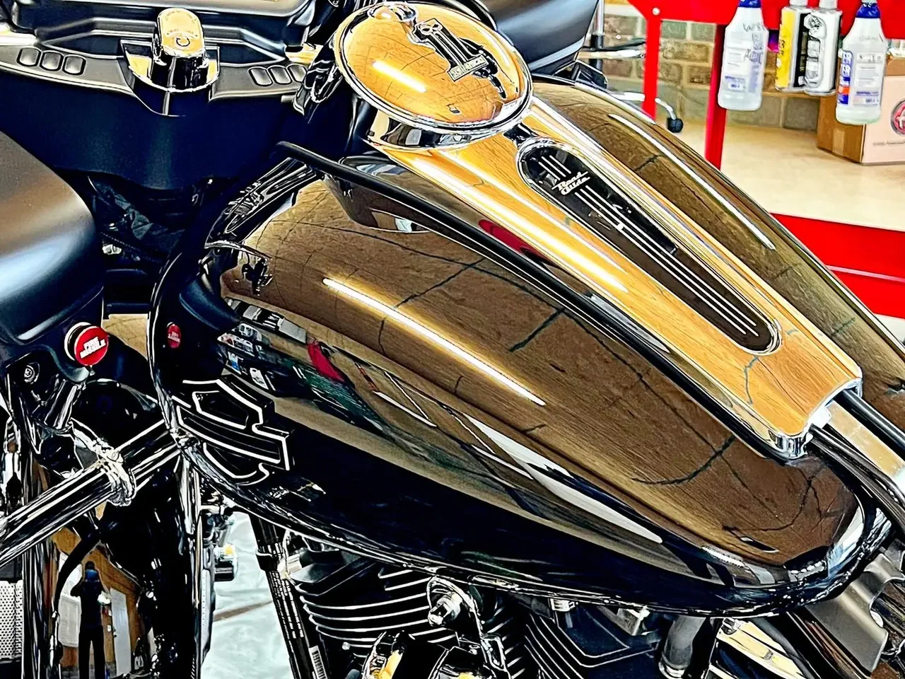 Close-up of a motorcycle's chrome fuel tank reflecting the workshop ceiling. The fuel cap features a logo, and the surrounding area includes parts of the handlebars and engine.
