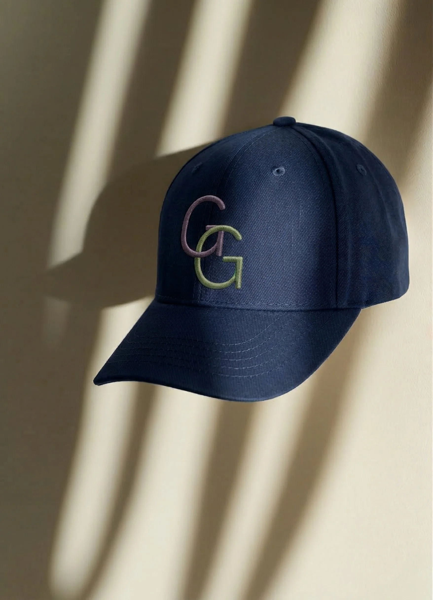A dark blue baseball cap with embroidered initials 'C' and 'G' hanging on a beige wall with shadows.
