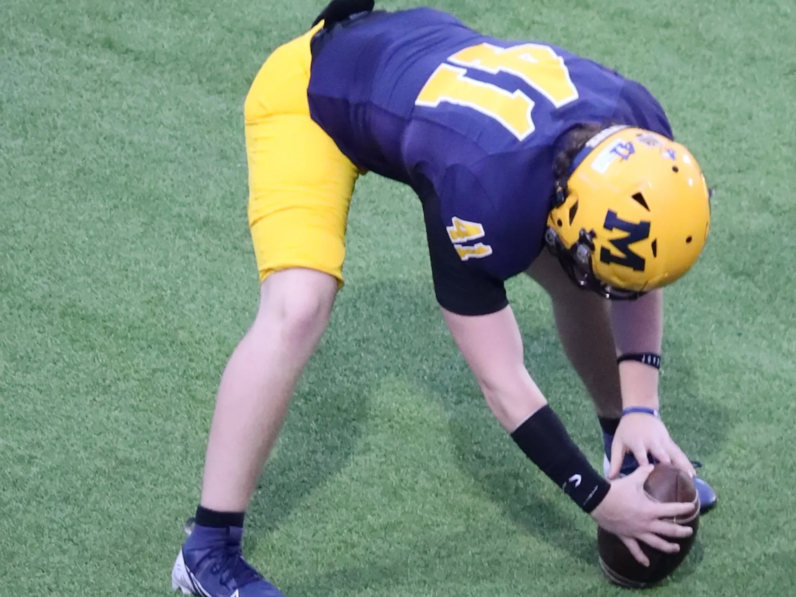A football player wearing a yellow helmet and a blue and yellow uniform, bending down and holding a football on a green field.