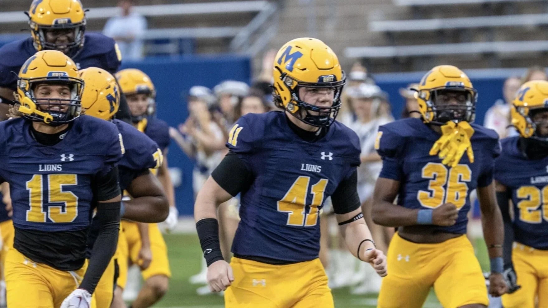 A group of high school football players in blue and yellow uniforms, wearing helmets, running on the field during a game.