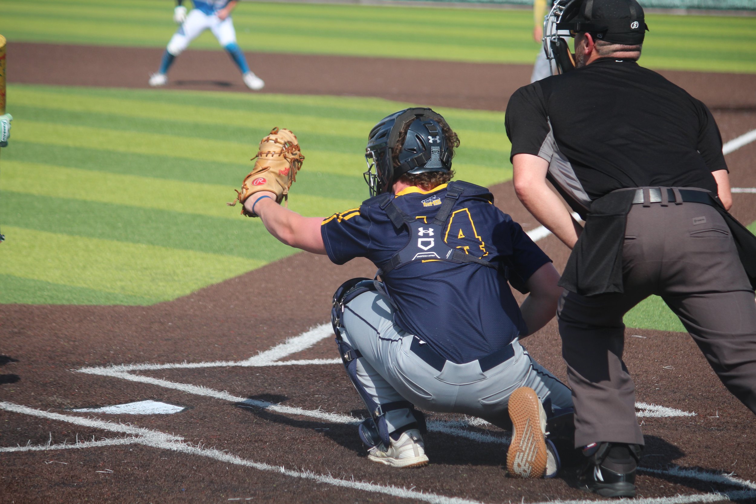 A baseball catcher crouching behind home plate in full gear with a mitt ready to catch a pitch, while an umpire stands beside him during a game on a baseball field.