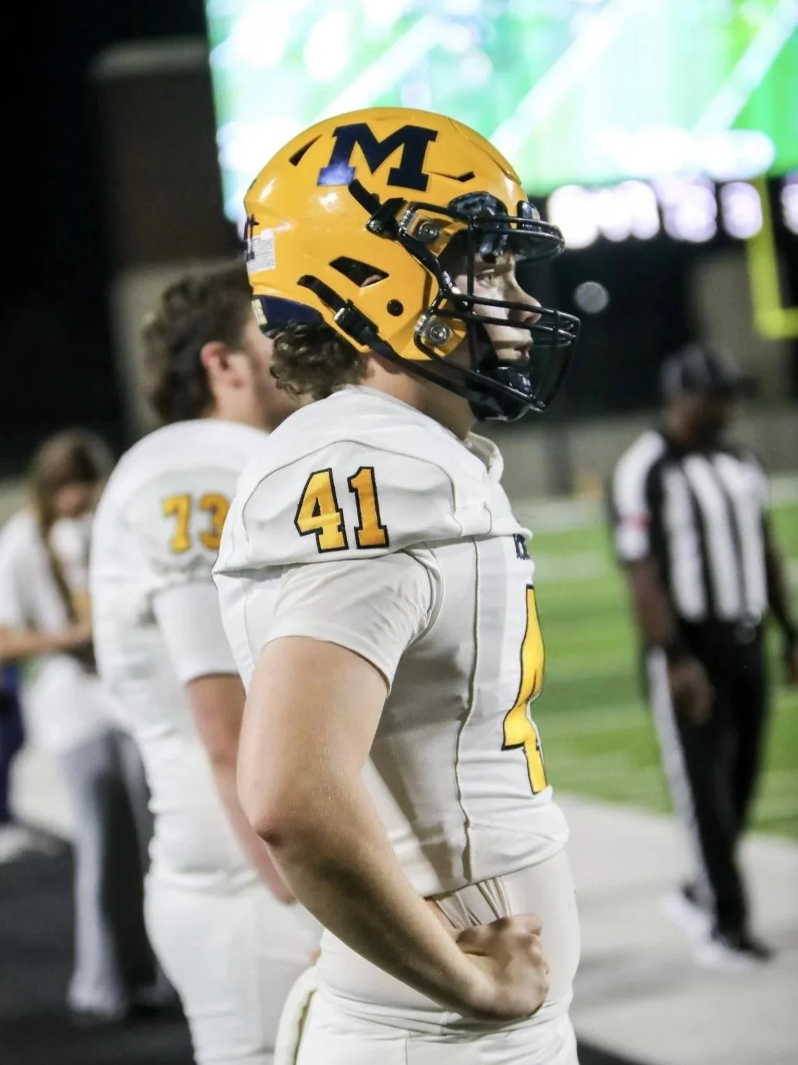 A football player wearing a yellow helmet with a black 'M' logo and a white jersey with the number 41 stands on the sideline of a football field. Other players and an official are in the background.