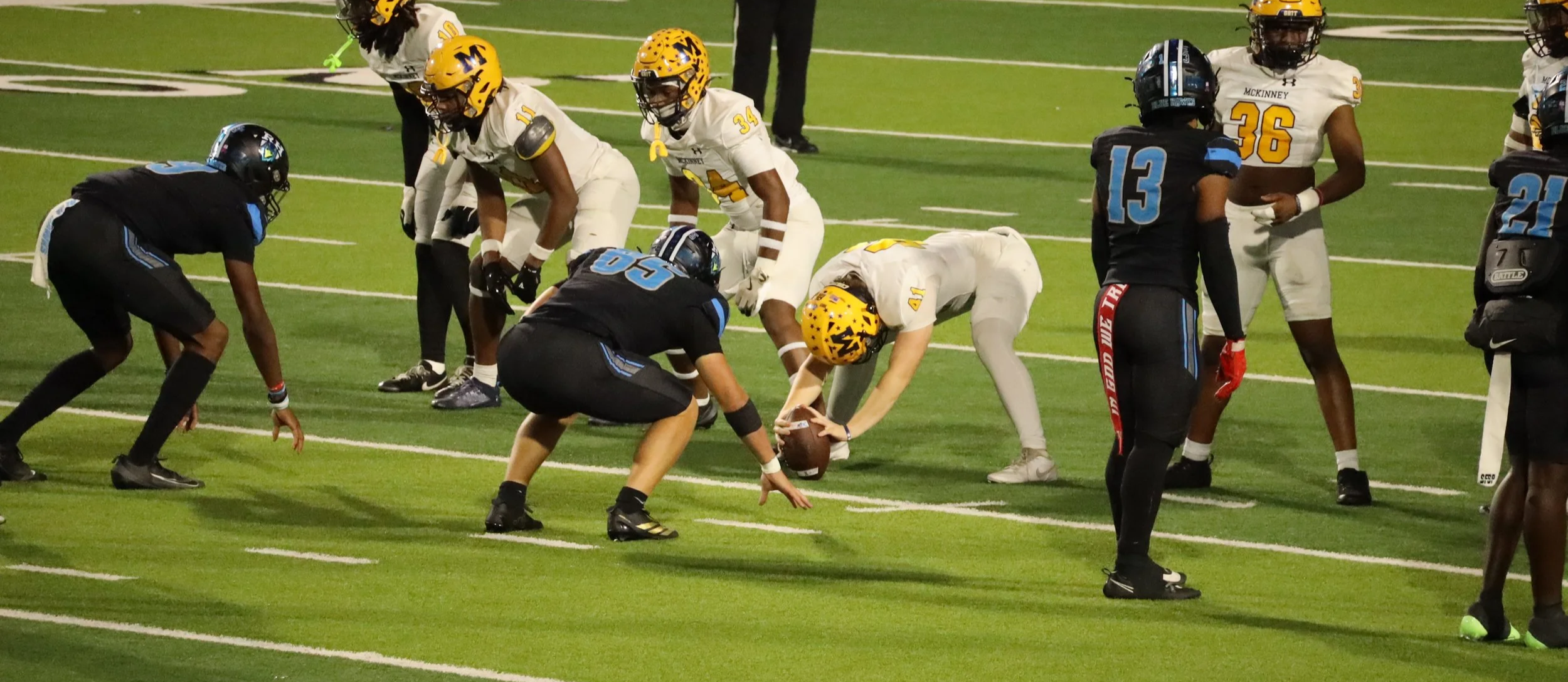 Football players from two teams lined up for a play on the field, with a player bending down to pick up the football as a teammate in white prepares for the snap.