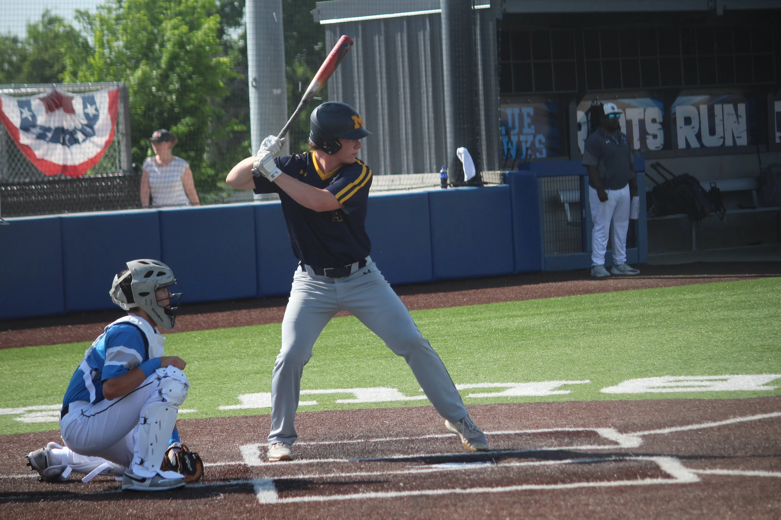 A baseball player stands at home plate, ready to bat, while a catcher crouches behind him. Both players are in uniform on a baseball field with a blue outfield wall and a few spectators in the background.