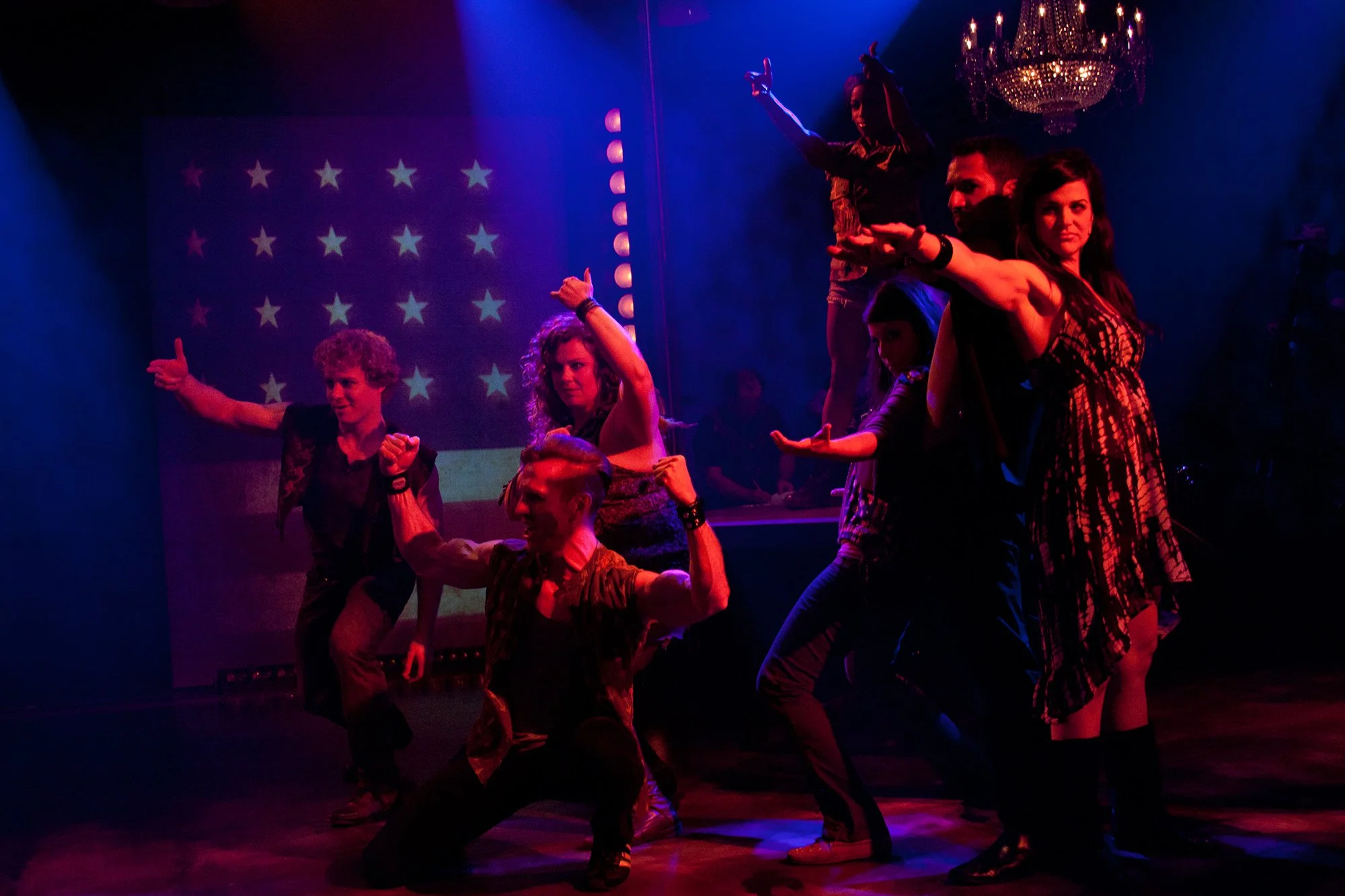 Group of performers on stage, some striking poses with arms extended, in front of a backdrop with stars; stage is lit with colorful lights and a chandelier hangs overhead.