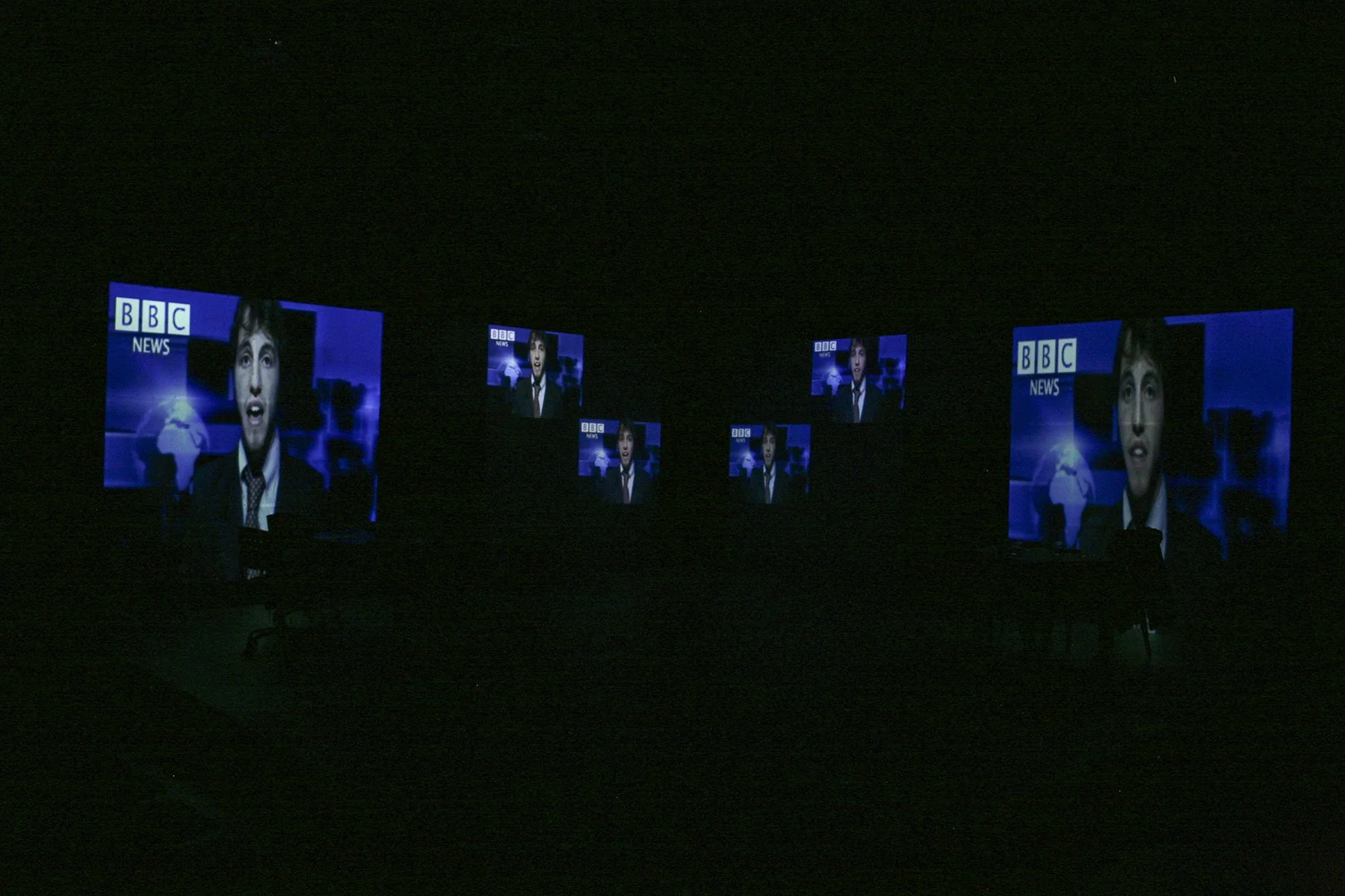 Multiple illuminated screens displaying a news anchor with short dark hair and a dark blazer, set against a blue background with the BBC News logo, in a dark room.