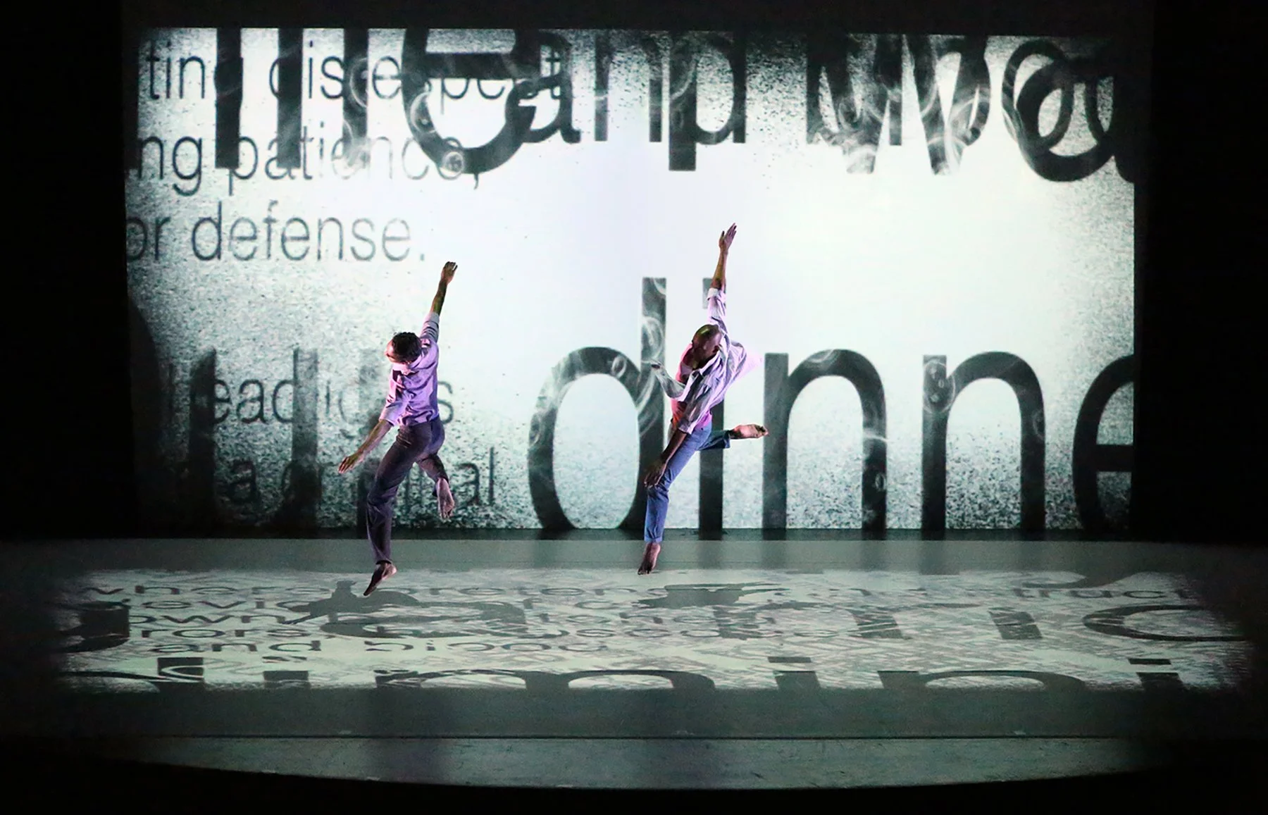Two dancers performing in front of a backdrop with text and projection effects.